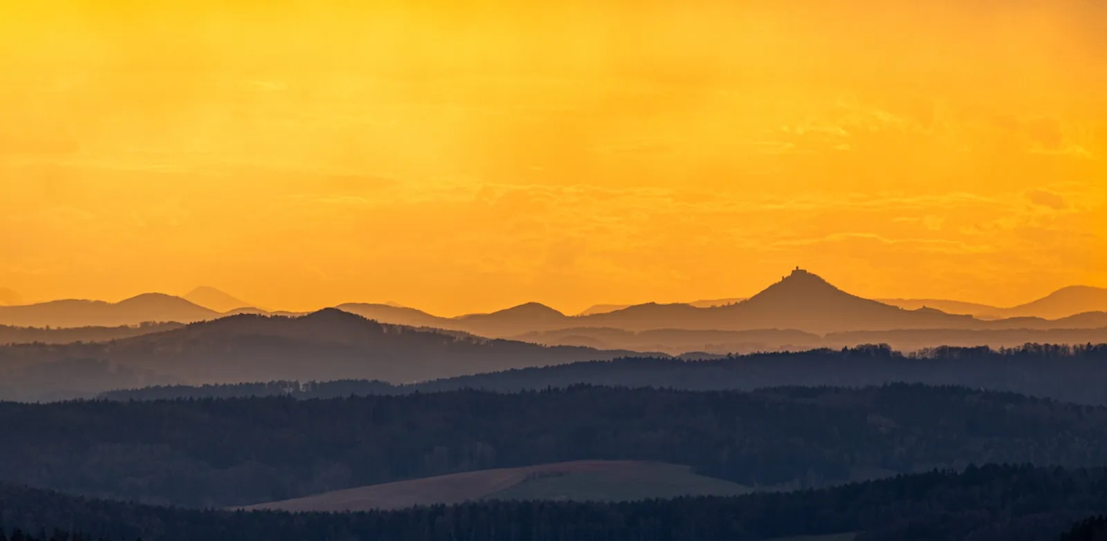 Bewaldete Alpengipfel unter einem leuchtenden goldenen Sonnenuntergang, schneebedeckte Grate durch dunstigen Nebel und neblige Täler hindurch sichtbar.