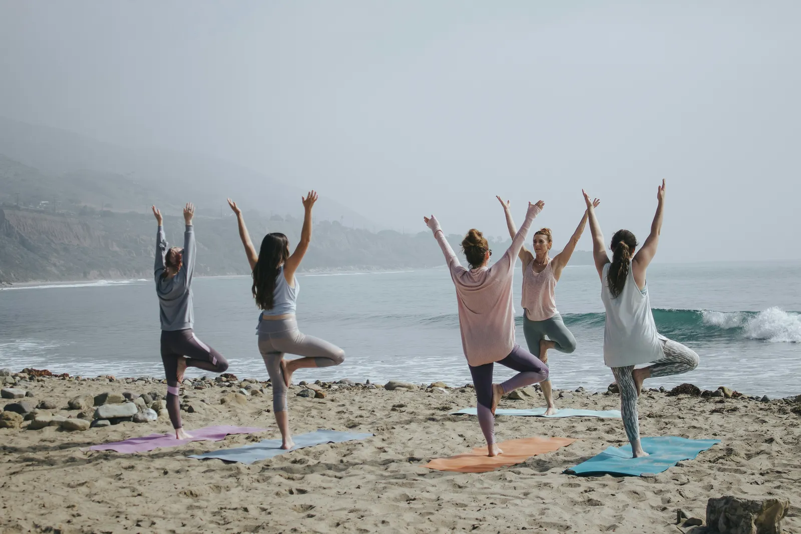 Gruppe von fünf Frauen in Baumpose auf Yogamatten an einem nebligen Strand mit Meeresklippen im Hintergrund