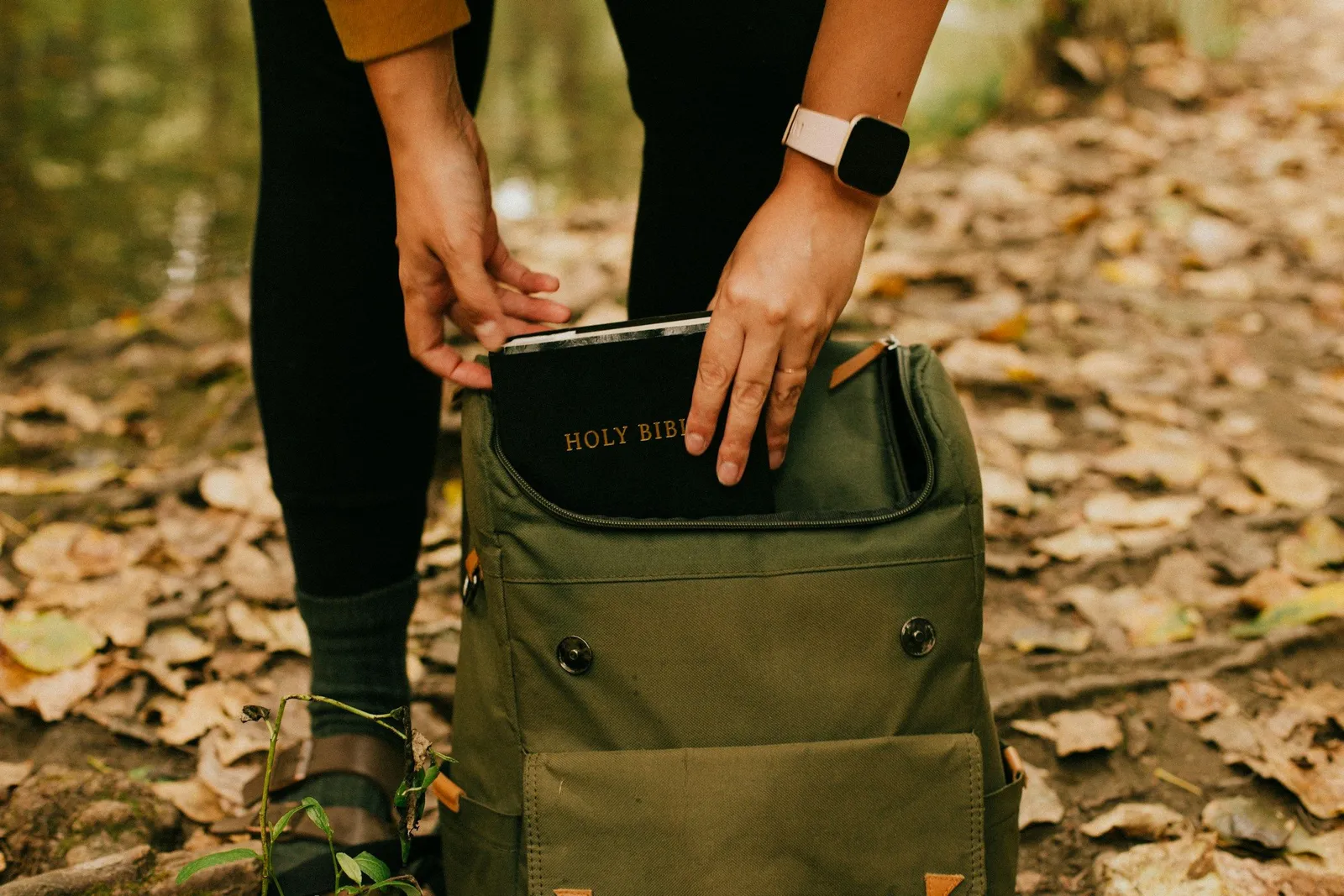 Person kauert in Herbstblättern und legt ein Buch in einen grünen Rucksack, Vorbereitung für eine spirituelle Auszeit im Freien