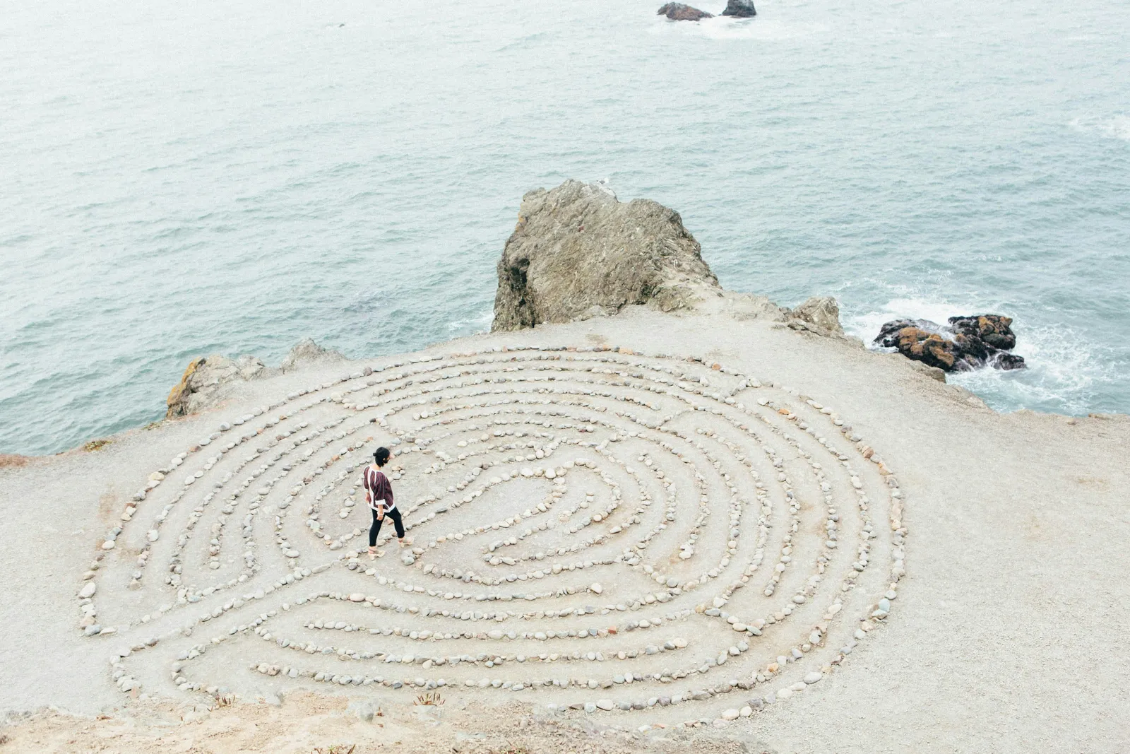 Vogelperspektive einer Person, die ein großes Stein-Labyrinth an einer Meeresklippe entlanggeht. Ein meditativer Weg ohne offensichtliche Abkürzung.