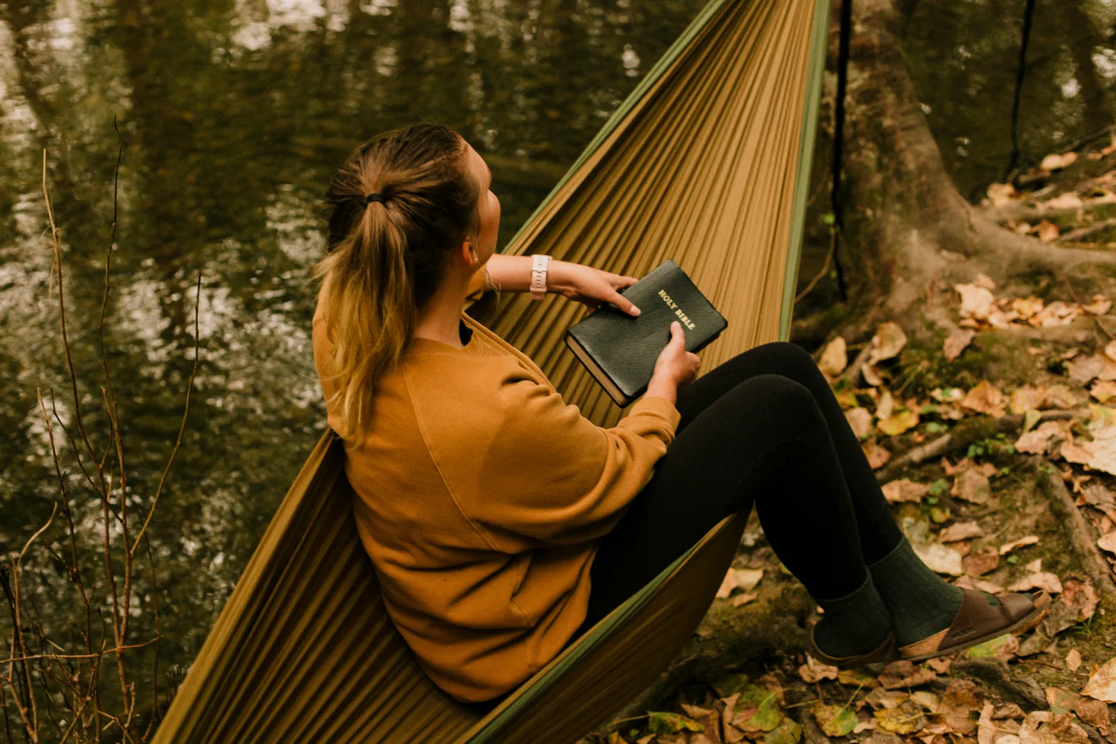 Frau in einem senfgelben Pullover, in einer grünen Hängematte an einem Waldteich sitzend und lesend.