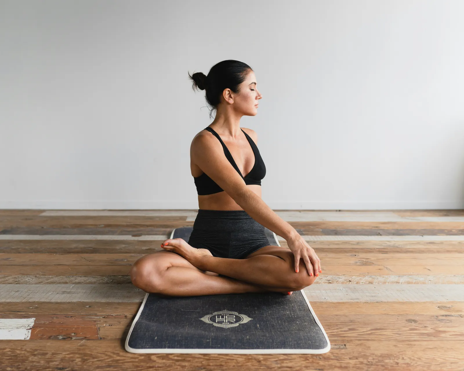 Frau in schwarzer Sportkleidung sitzt mit geschlossenen Augen im Lotus-Sitz auf einer Yogamatte auf einem Holzboden.