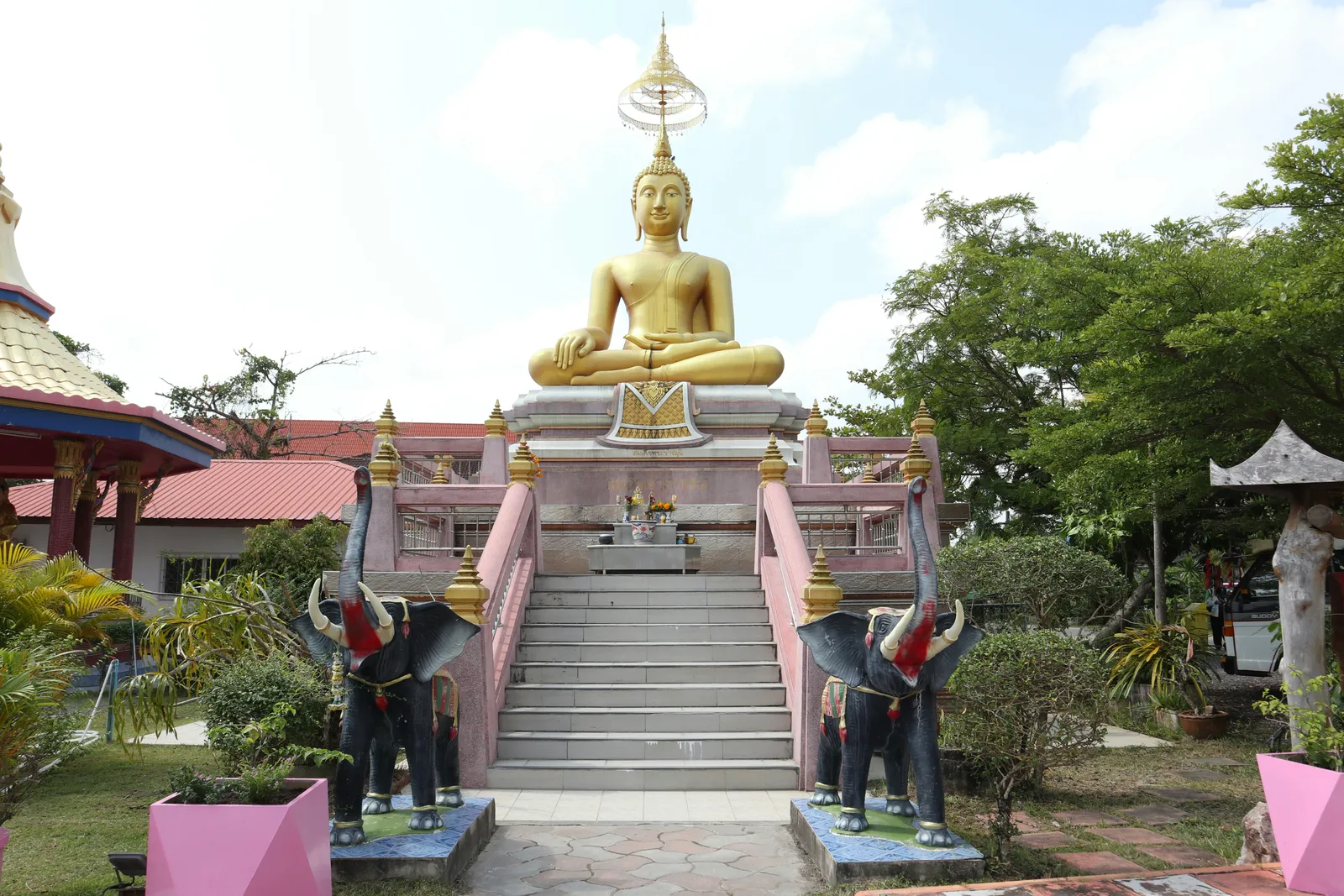 Große goldene Buddha-Statue auf einem erhöhten Sockel, flankiert von Elefantenskulpturen an einem Thai-Tempel im hellen Tageslicht.