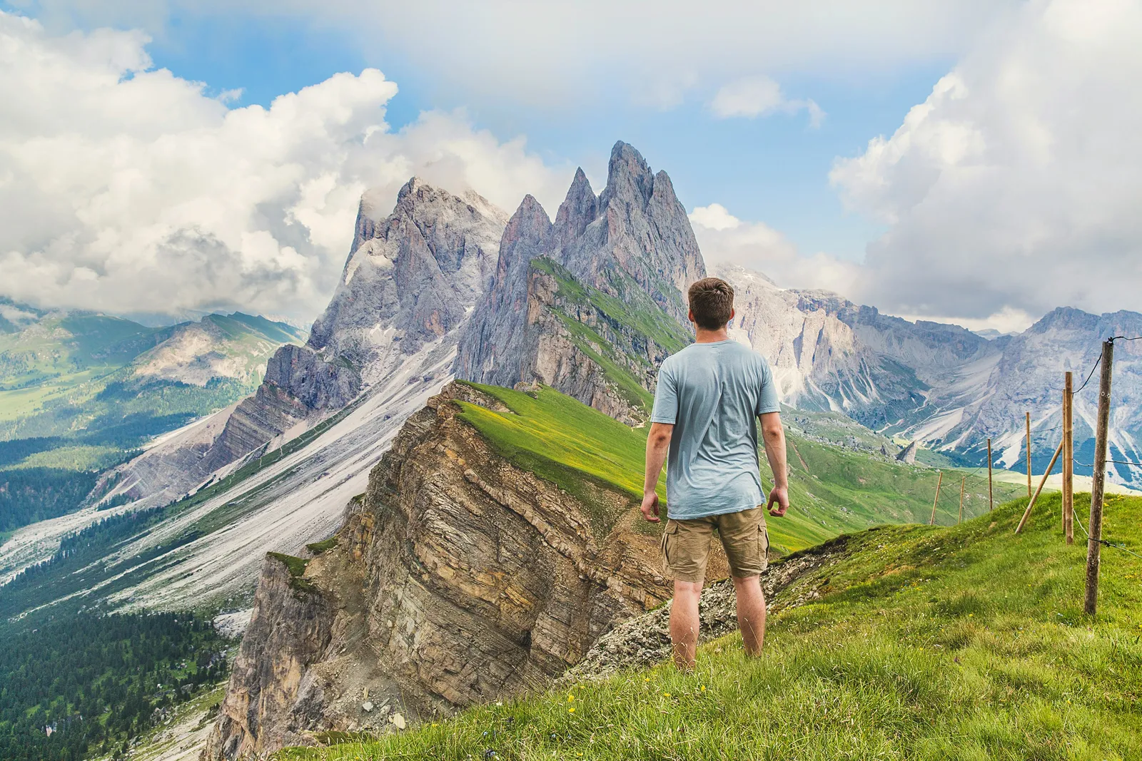Ein Mann mit Rucksack steht auf einem grasbewachsenen Gebirgsrücken und blickt auf zackige Bergspitzen unter bewölktem Himmel.