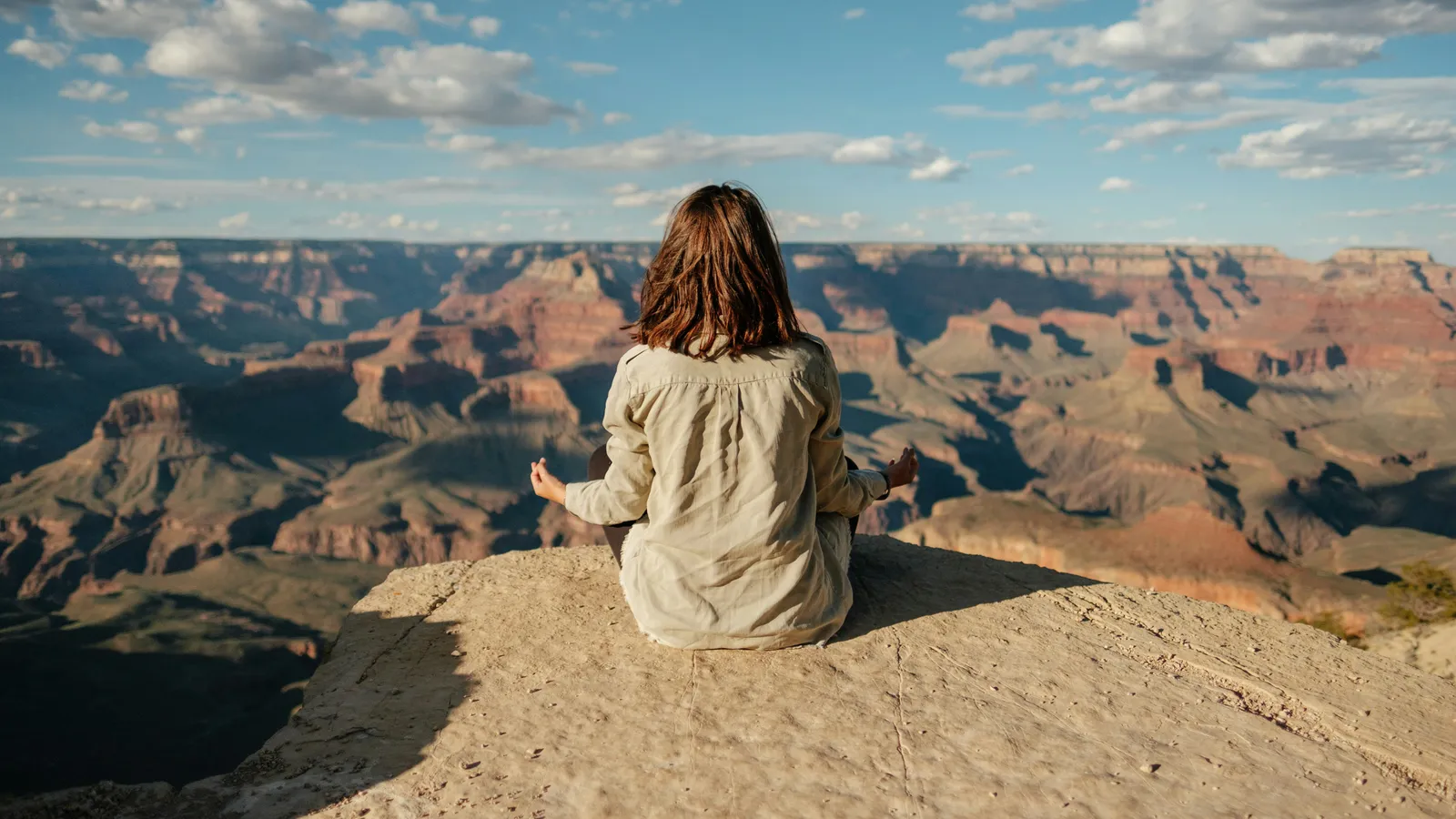 Frau in einem beigen Leinenhemd sitzt im Schneidersitz auf einem Sandsteinvorsprung am Rand eines Canyons, Hände in Meditationsmudra, Blick über geschichtete rote Felsformationen.