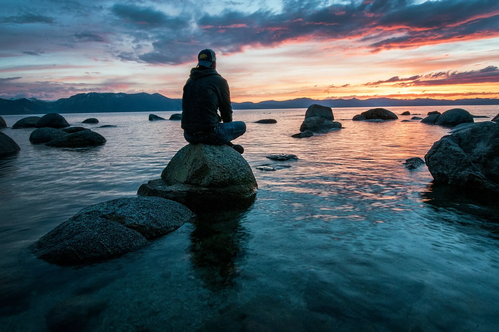 Person in einer dunklen Kapuzenjacke sitzt im Schneidersitz auf einem Felsen in einem stillen See bei Sonnenuntergang, dramatische Sturmwolken am Himmel