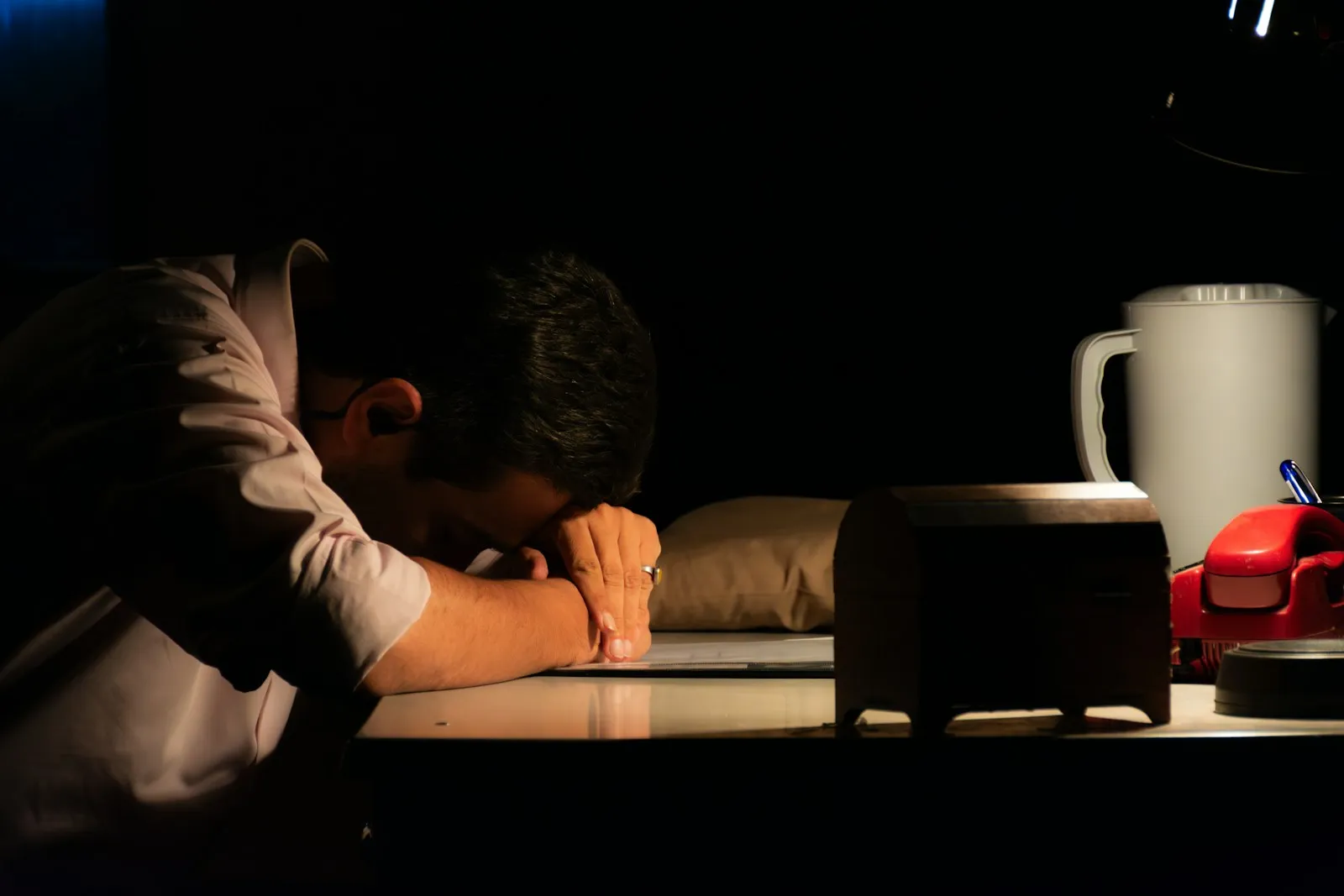 Eine Person in dunkler Kleidung, Kopf nachts bei Kerzenlicht auf dem Schreibtisch, starke Schatten neben einem Laptop.