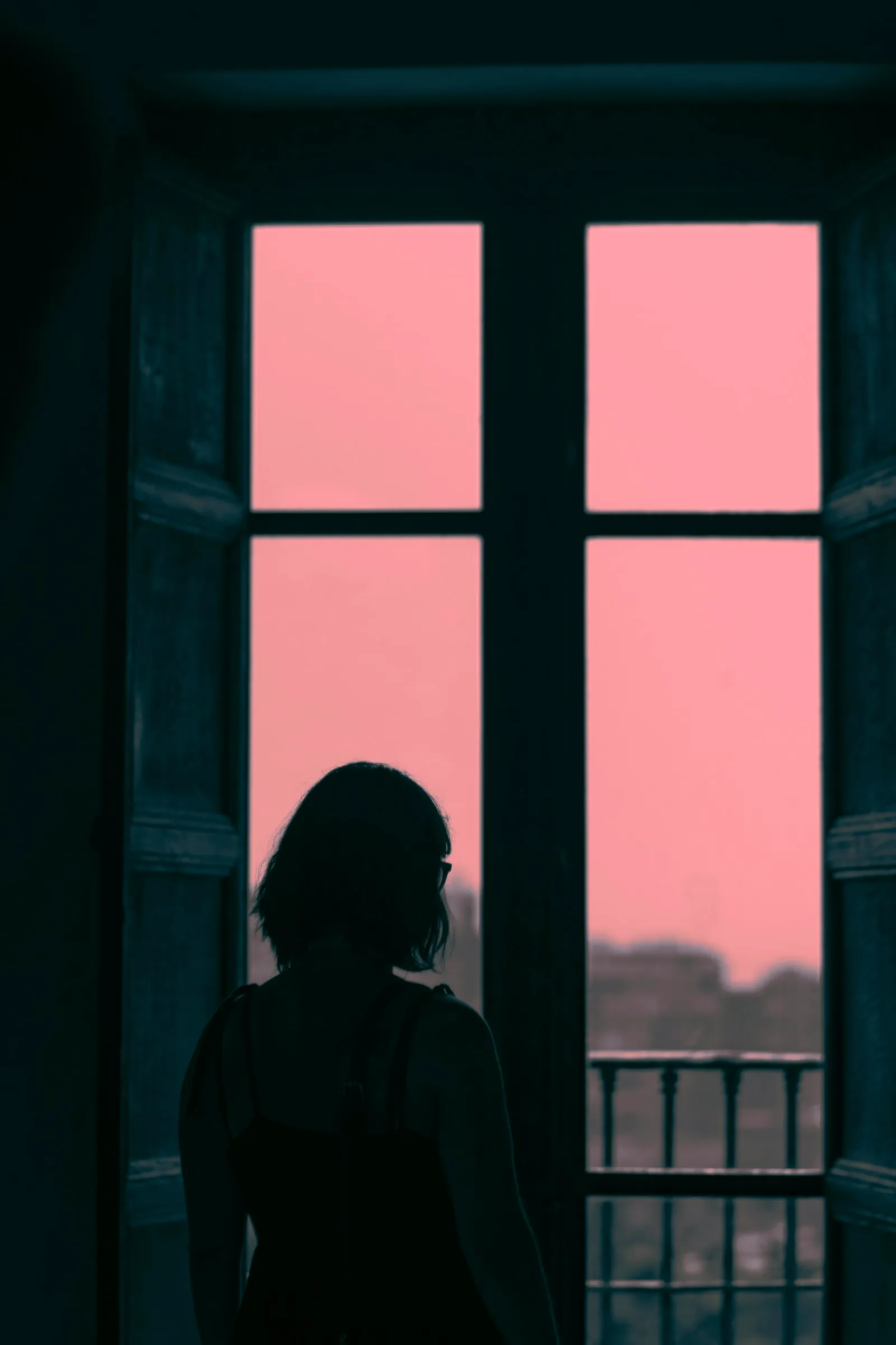 Silhouette einer Person, die an einem großen Sprossenfenster steht, im Gegenlicht vor einem lachsfarbenen Abendhimmel.