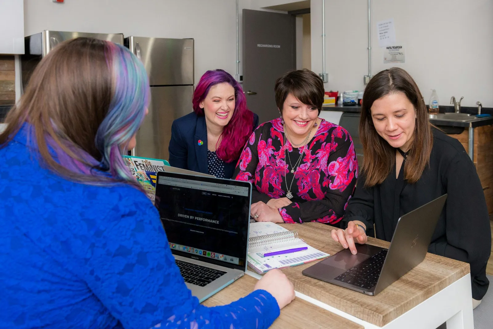 Vier Frauen arbeiten gemeinsam glücklich an einem Holztisch mit zwei Laptops in einer hellen Büroküche. Lockere, energetische Workshop-Atmosphäre.