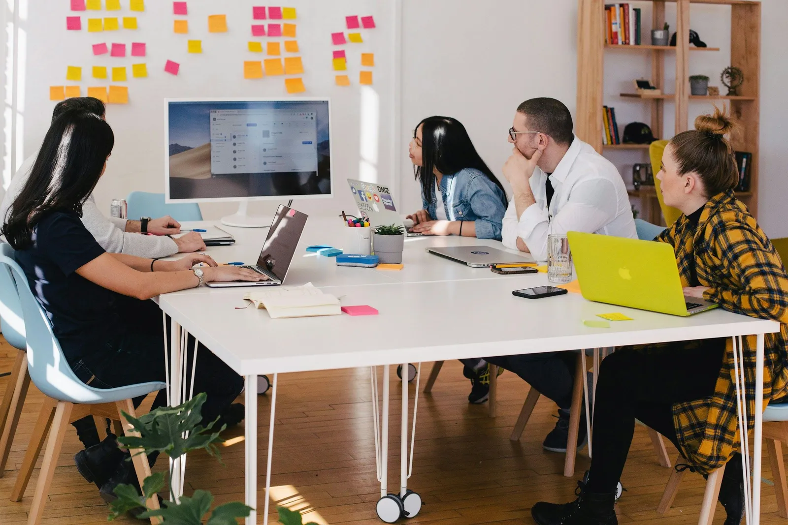 Fünf Personen arbeiten gemeinsam an einem weißen Tisch mit Laptops und Notizblöcken in einem hellen Büro mit bunten Haftnotizen an der Wand. Team-Brainstorming-Meeting.