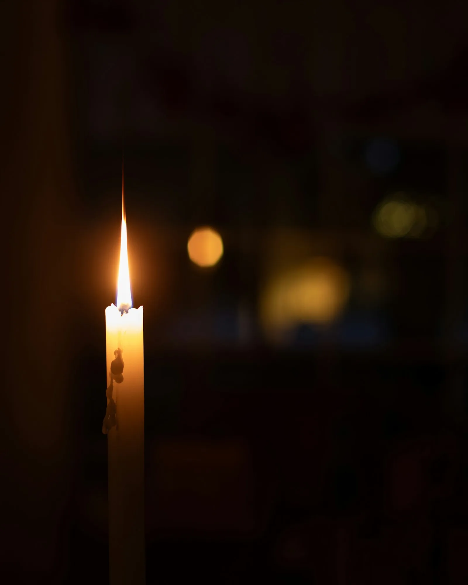 Eine schlanke weiße Kerze brennt vor einem dunklen Raum. Im unscharfen Hintergrund leuchten einzelne warme Lichtpunkte.
