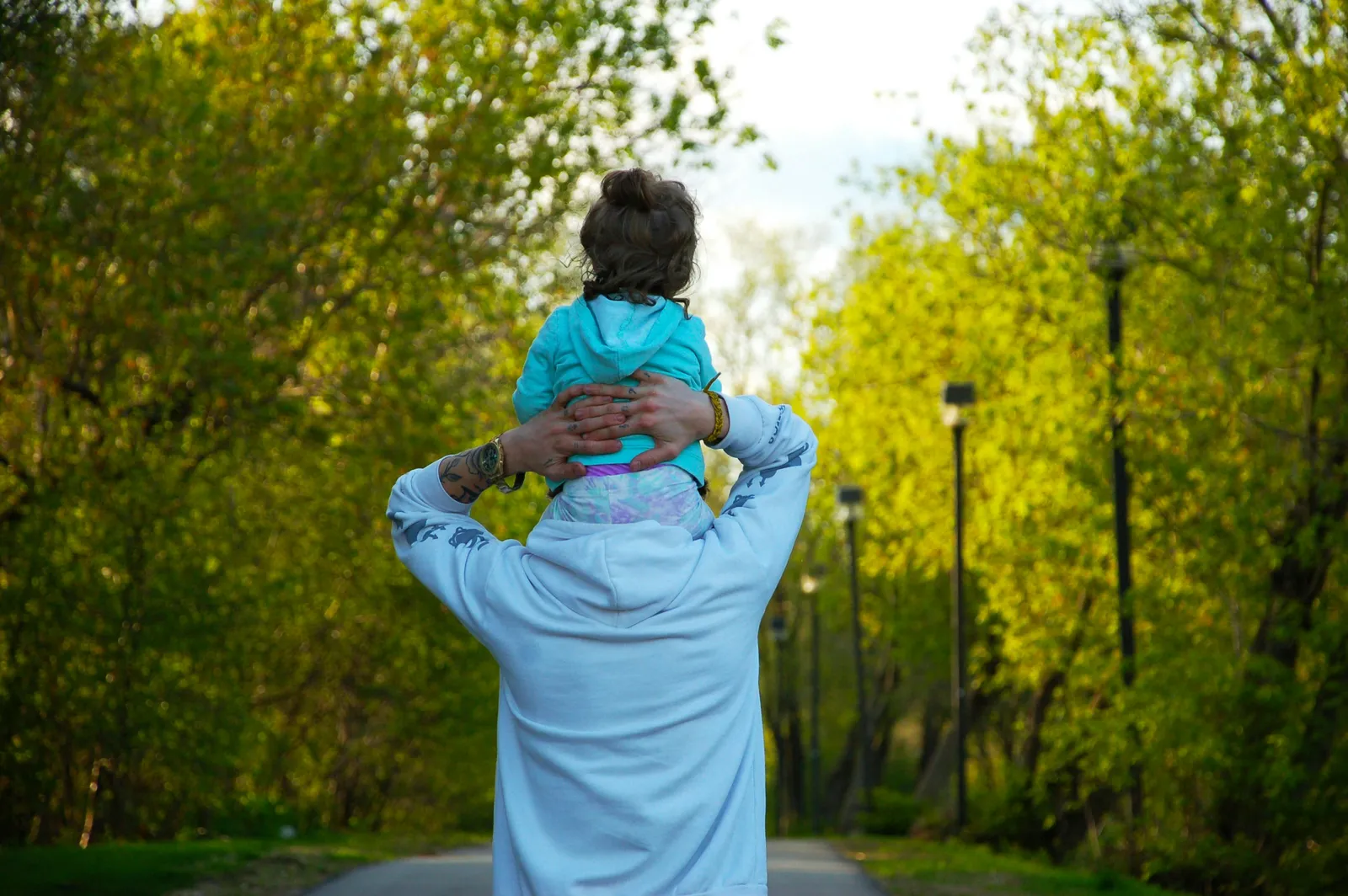 Ein Erwachsener geht von hinten gesehen eine grüne Allee mit Laternen entlang und trägt ein kleines Kind im türkisen Kapuzenpulli auf den Schultern.
