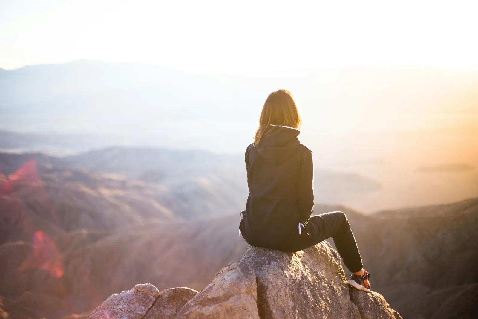 Frau in schwarzer Kapuzenjacke blickt auf nebelbedeckte Bergketten bei Sonnenaufgang von einem Felsvorsprung, warmes Licht im Hintergrund.