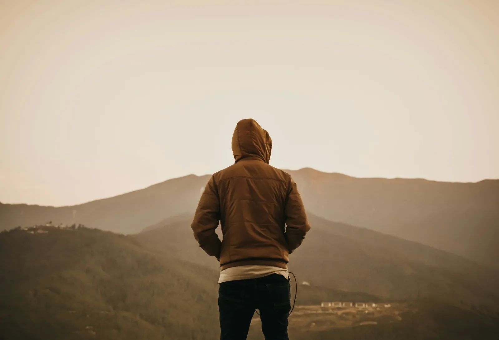 Ein Mann mit Kapuzenjacke steht mit dem Rücken zur Kamera und blickt auf nebelverhangene Berge in der Ferne. Sepia-Töne, introspektiv.