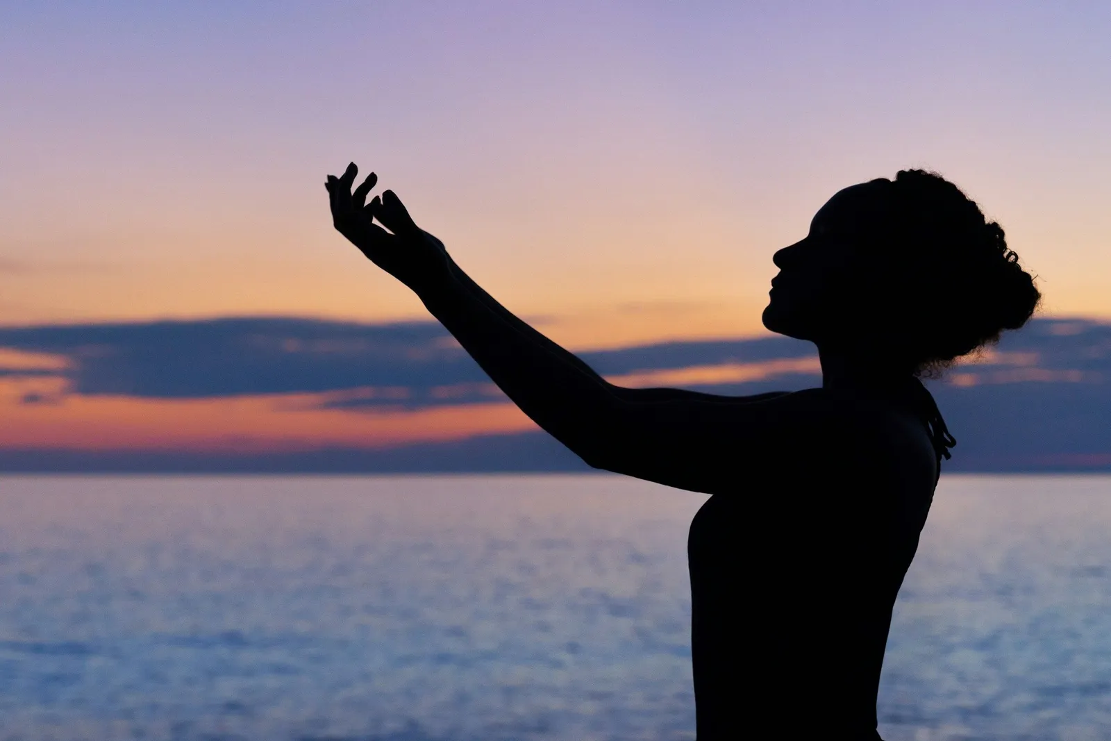 Silhouette einer Frau, die einen Arm anmutig gegen einen weichen, pastell-violetten, rosa und orangefarbenen Sonnenuntergangshimmel über ruhigem Wasser streckt. Stille, ehrfürchtige Abenddämmerungsszene.