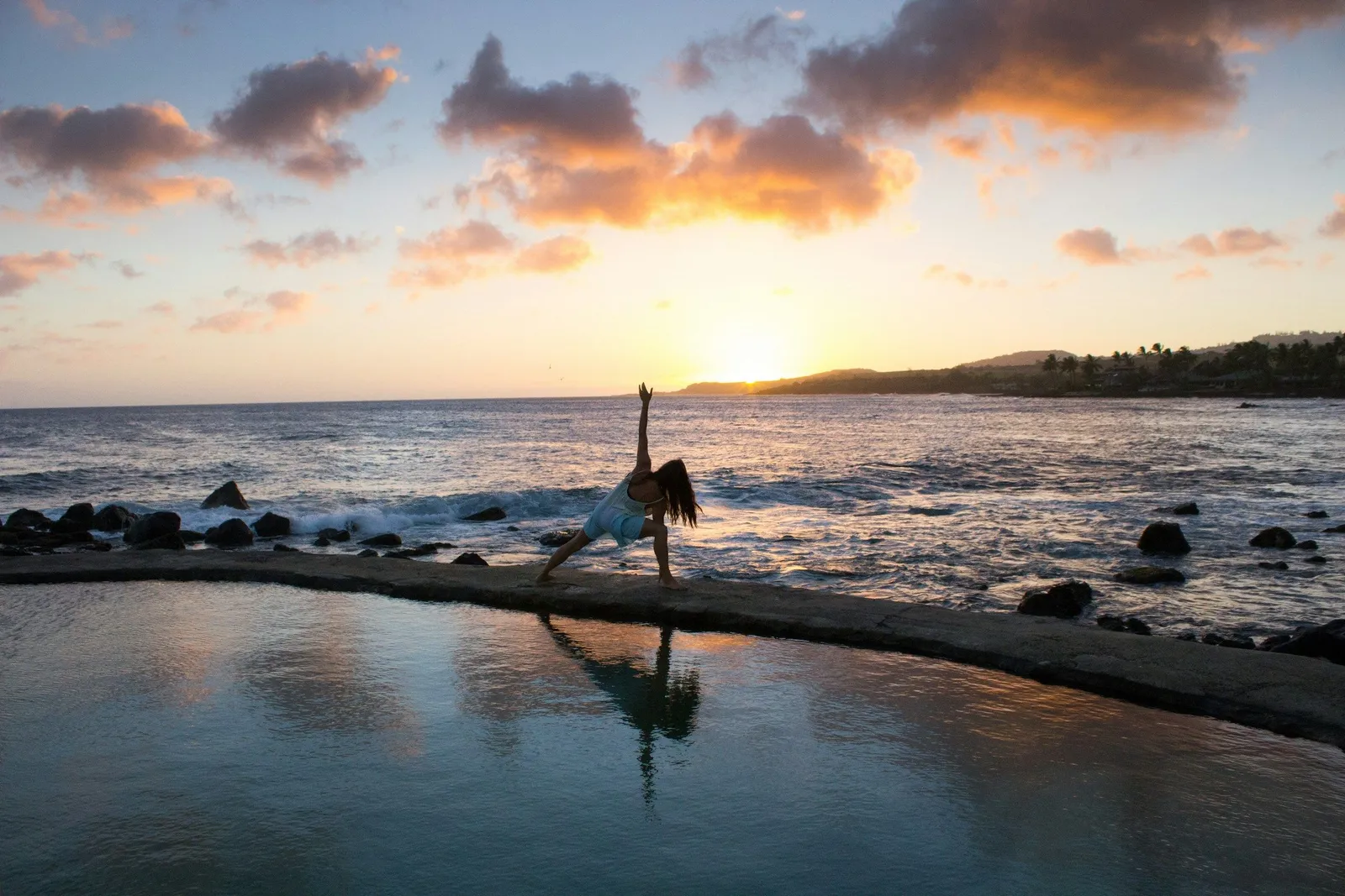 Frau in einer Yoga-Seitenbiegung auf einer Steinterrasse neben einem Infinity-Pool bei Sonnenuntergang.