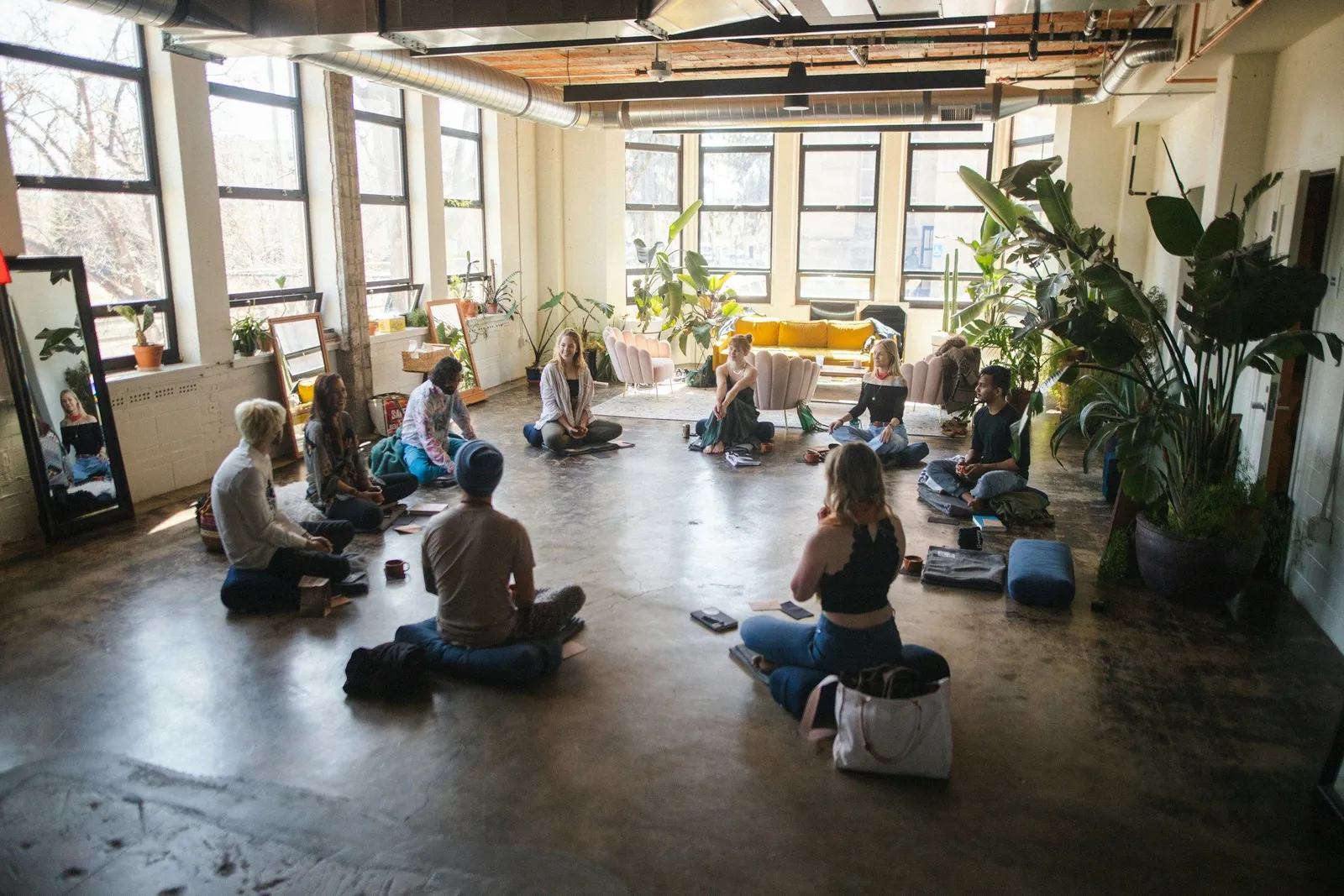 Gruppe von Menschen sitzt im Kreis auf dem Boden eines sonnenüberfluteten Loft-Studios mit Pflanzen, in einer Workshop- oder Gruppenrunde.