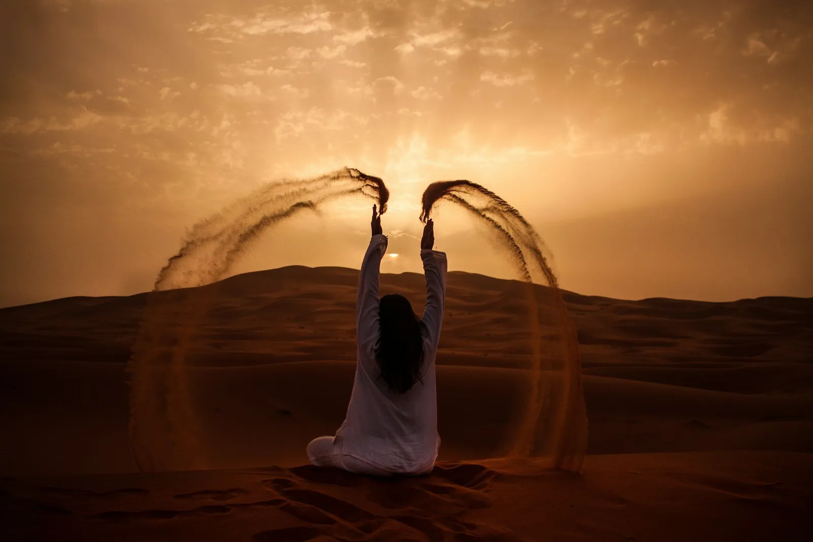 Silhouette einer Person in Weiß, die in der Wüste bei Sonnenuntergang sitzt, Arme erhoben, zwei Bögen Sand in den orangefarbenen Himmel wirft.