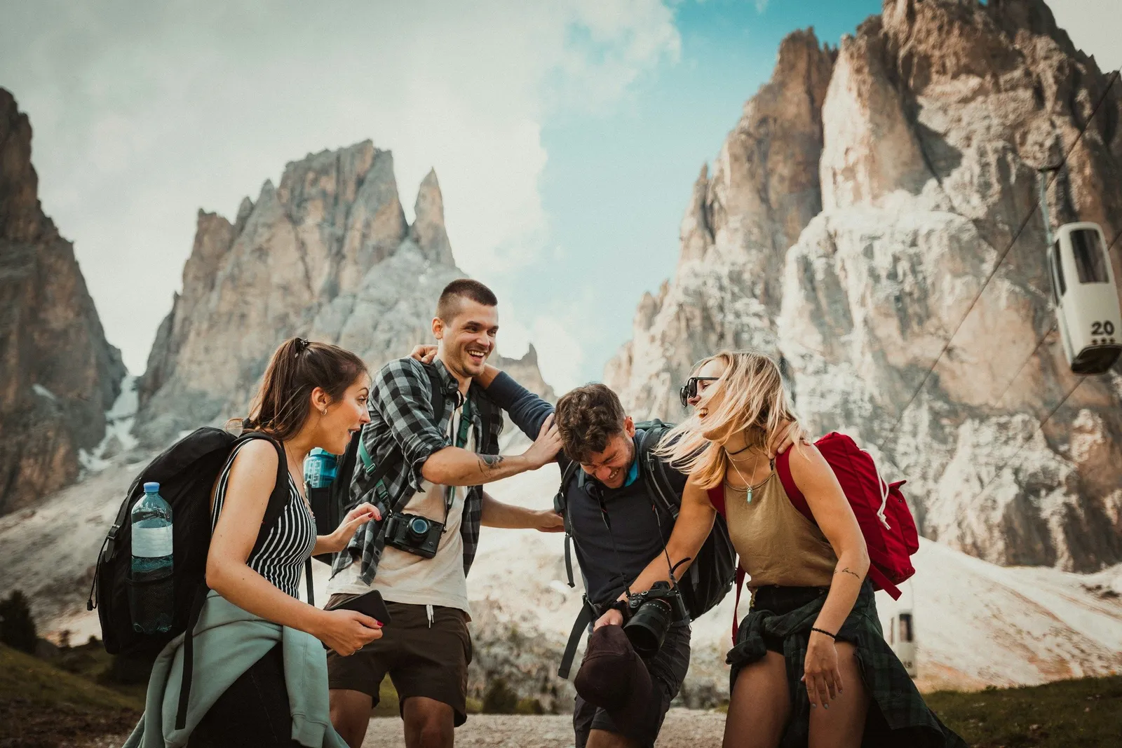Vier Freunde mit Rucksäcken lachen beim Wandern in den Dolomiten, dramatische Felsgipfel im Hintergrund. Aktiv und freudig.