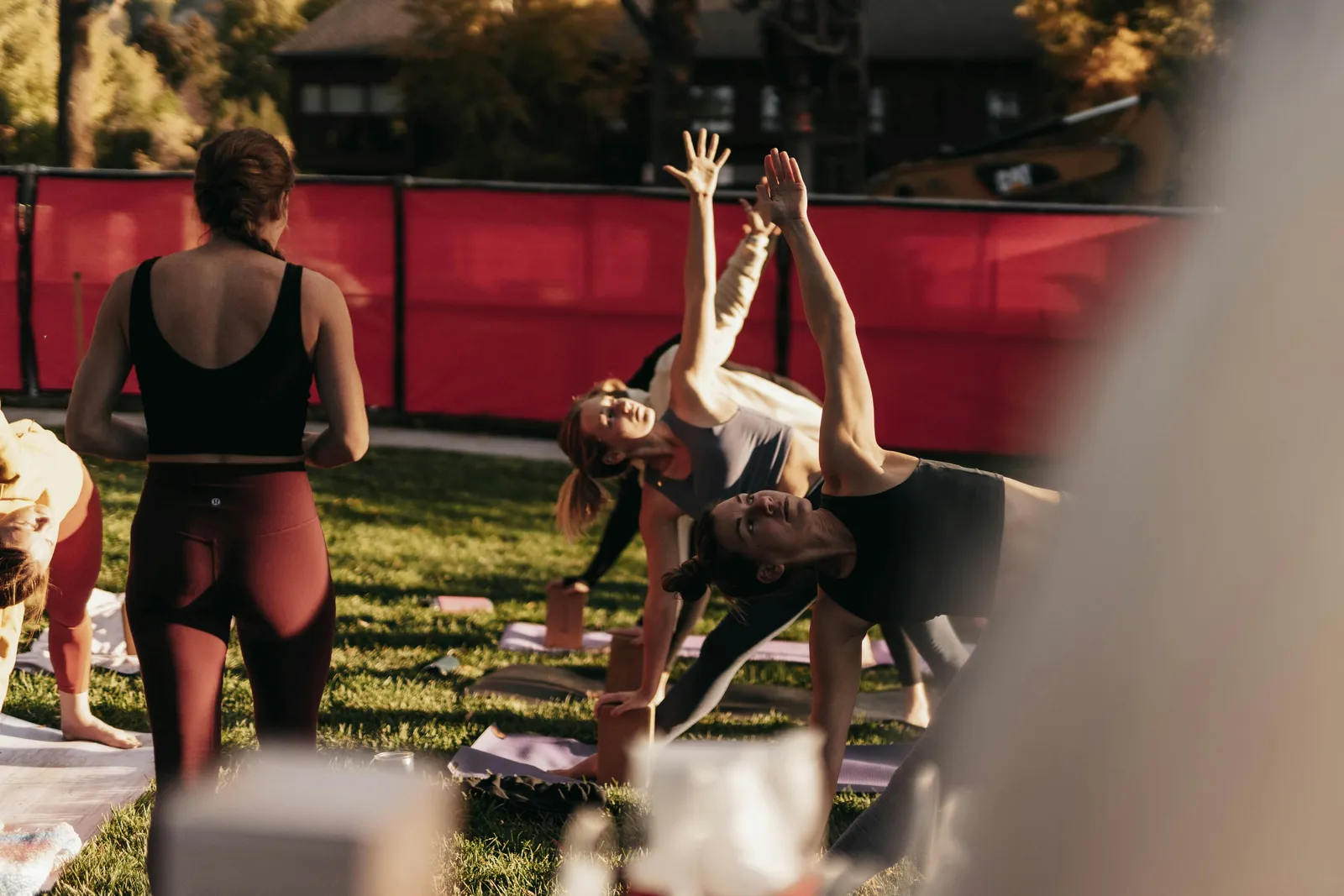 Frauen üben Dreieckshaltung auf Yogamatten auf einer Wiese im warmen Nachmittagslicht, roter Zaun im Hintergrund. Outdoor-Gruppenyo ga auf einem Retreat.