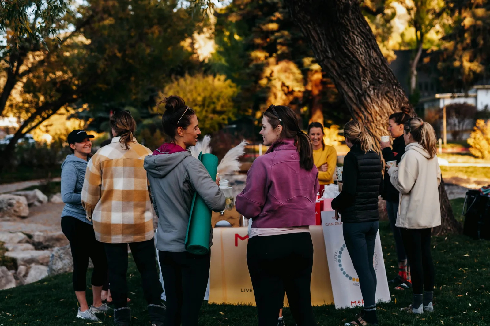 Gruppe Frauen versammelt sich um einen Gemeinschaftstisch im Freien in warmem Herbstlicht, Yogamatten und Taschen in der Hand. Austausch vor Retreat-Beginn.