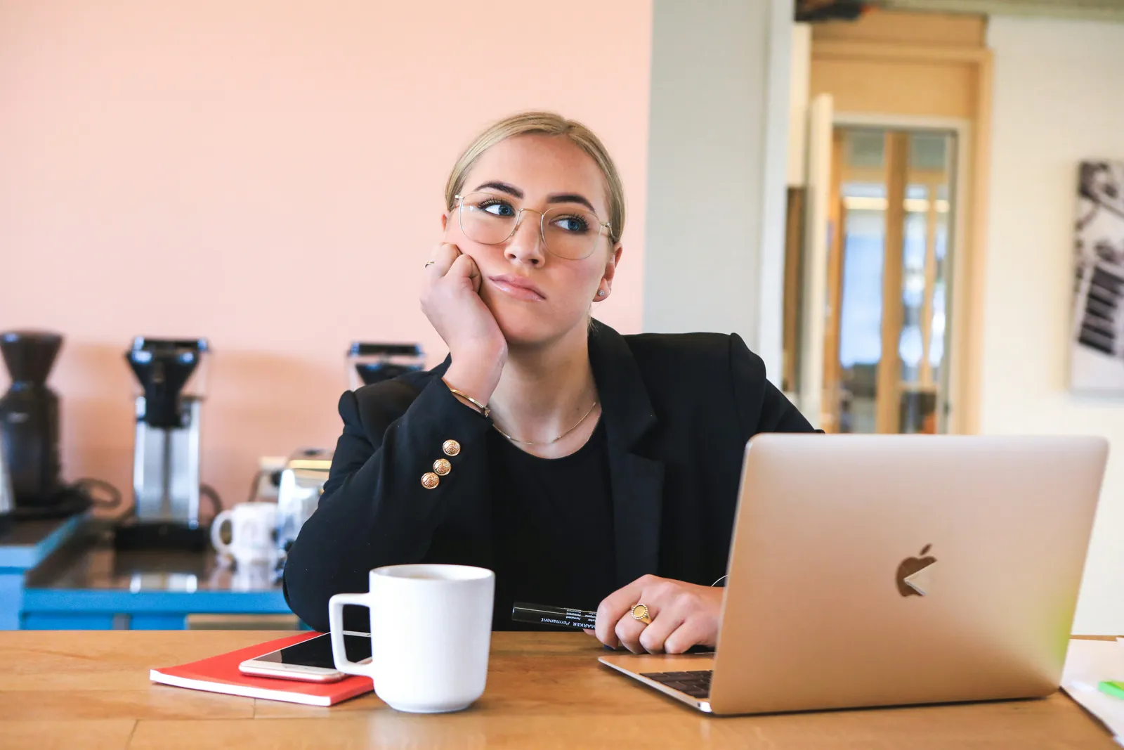 Frau in schwarzem Blazer lehnt den Kopf auf die Hand und schaut abwesend zur Seite, in einem Café mit Laptop und Kaffee. Erschöpfung vor der Auszeit.