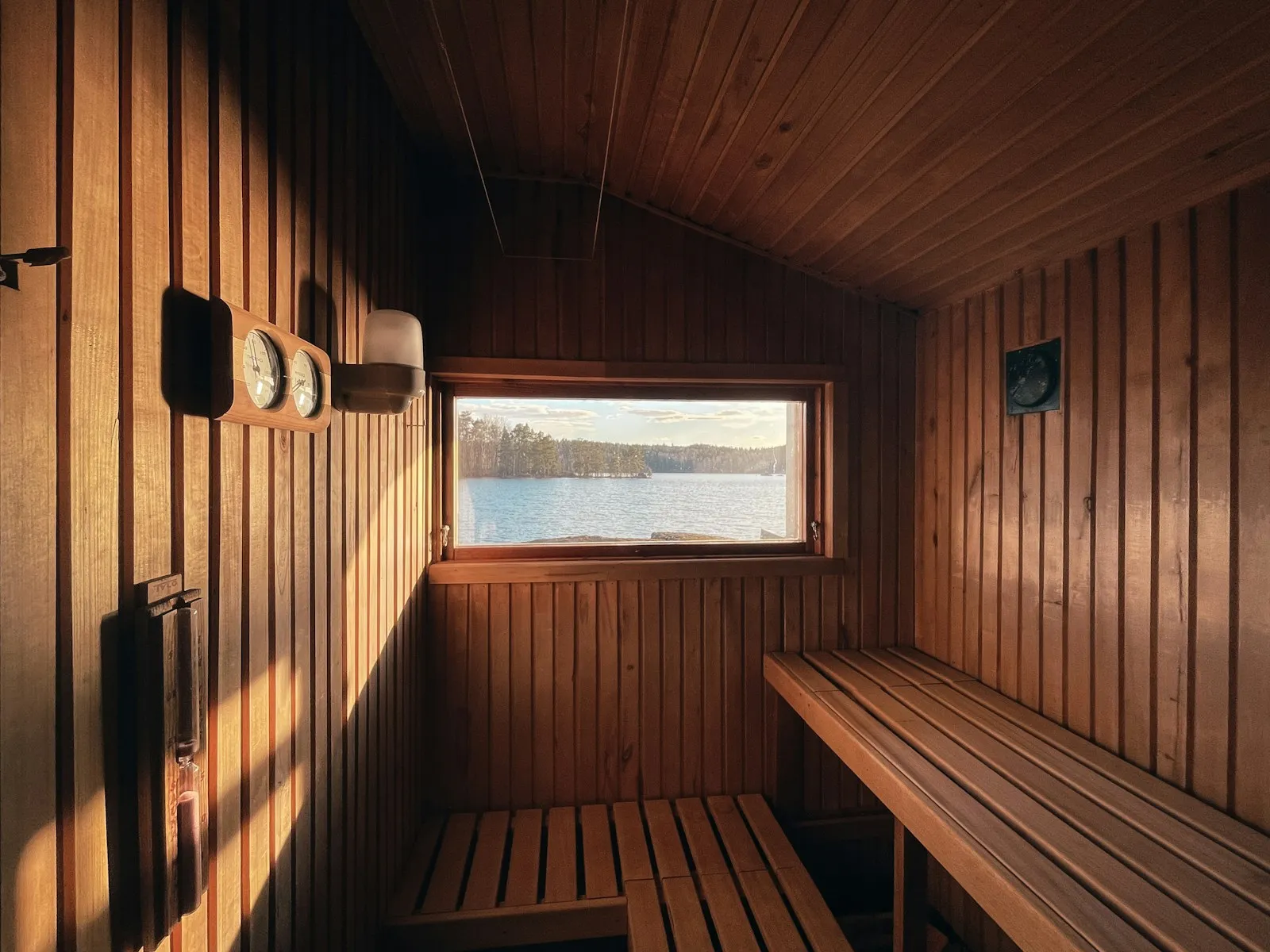 Saunainnenraum mit Holzbänken und großem Fenster, durch das ein See und ein Wald im warmen Tageslicht zu sehen sind