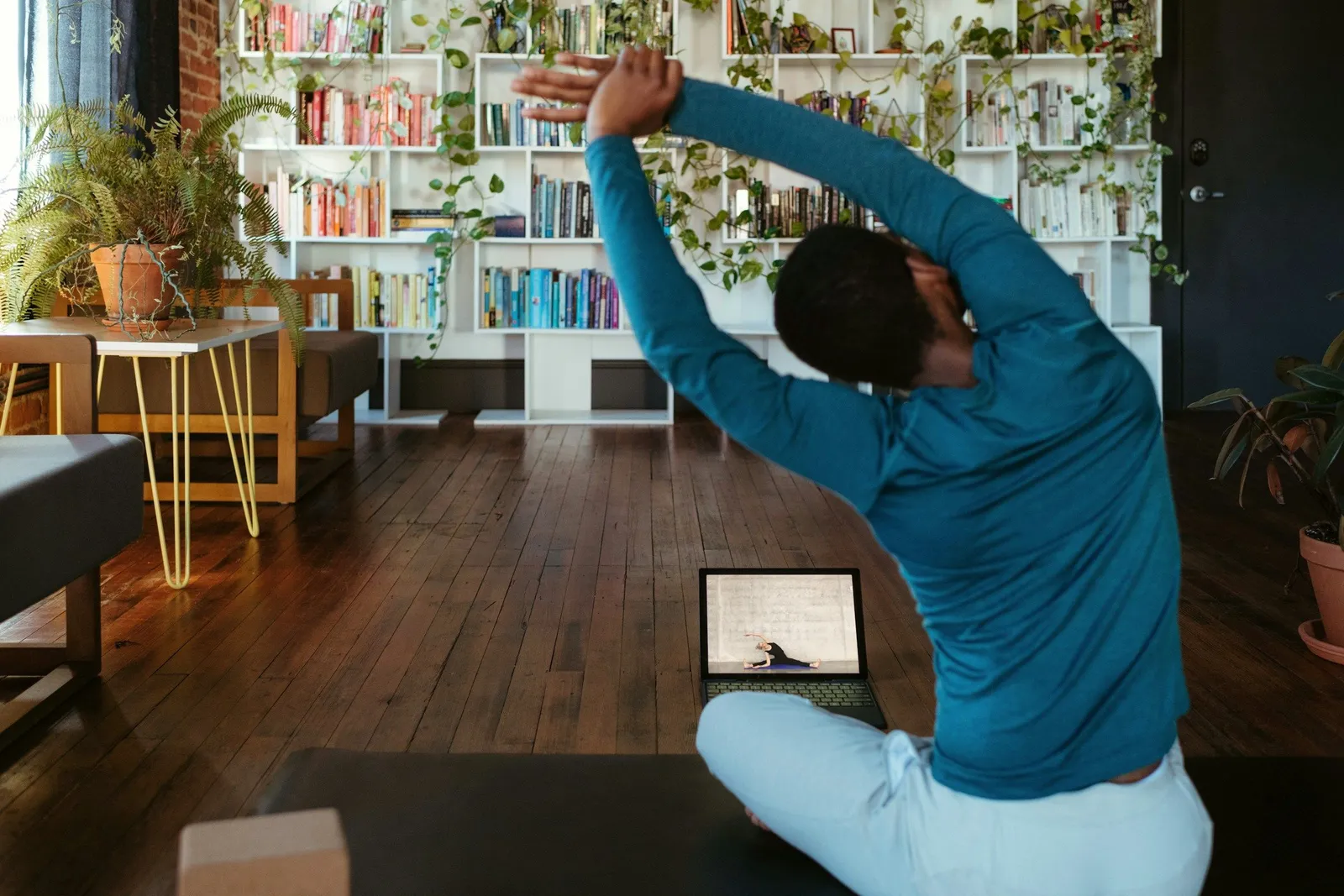 Ein Mann sitzt im Schneidersitz auf einer Matte und dehnt sich seitlich mit erhobenen, verschränkten Armen, vor sich ein Laptop mit einer Online-Yoga-Einheit, im Hintergrund ein Bücherregal mit Pflanzen.