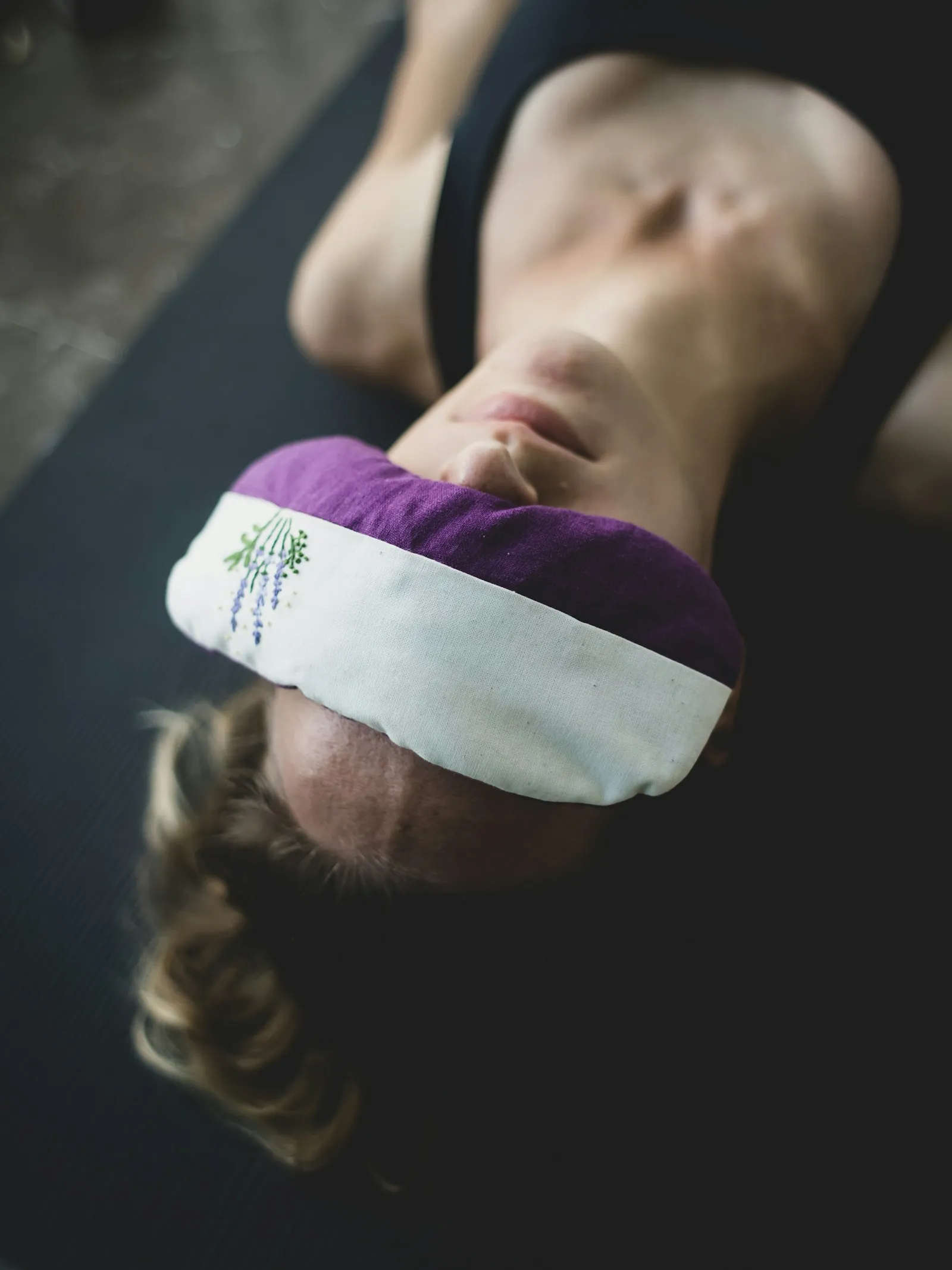 Eine Frau liegt auf einer dunklen Yogamatte auf dem Rücken, ein lila-weißes Augenkissen mit gestickten Lavendelzweigen bedeckt ihre Augen, das Licht ist gedämpft.