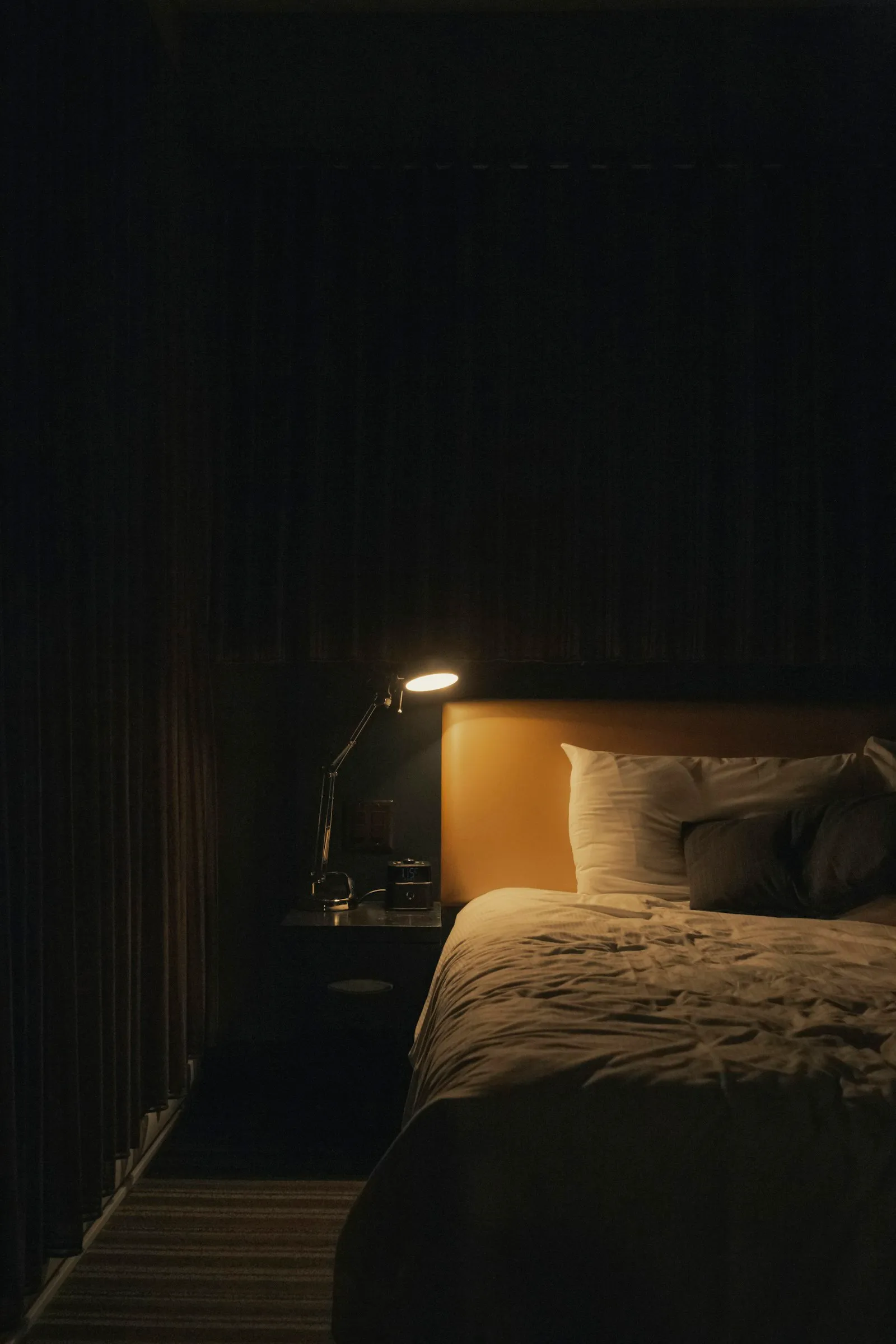 Ein dunkles Schlafzimmer, eine Gelenk-Leselampe auf dem Nachttisch wirft warmes Licht auf das Kopfteil und weiße Kissen eines ungemachten Bettes, links fallen schwere Vorhänge.
