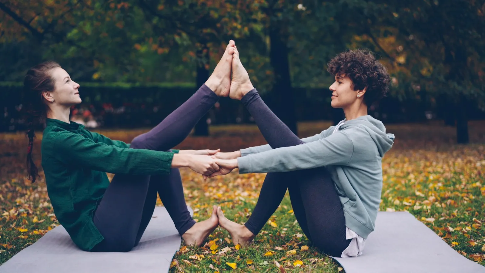 Zwei Frauen sitzen sich im Herbstpark auf Matten gegenüber, halten sich an den Händen und strecken je ein Bein nach oben, die Fußsohlen liegen oben aneinander, die anderen Füße stehen am Boden.