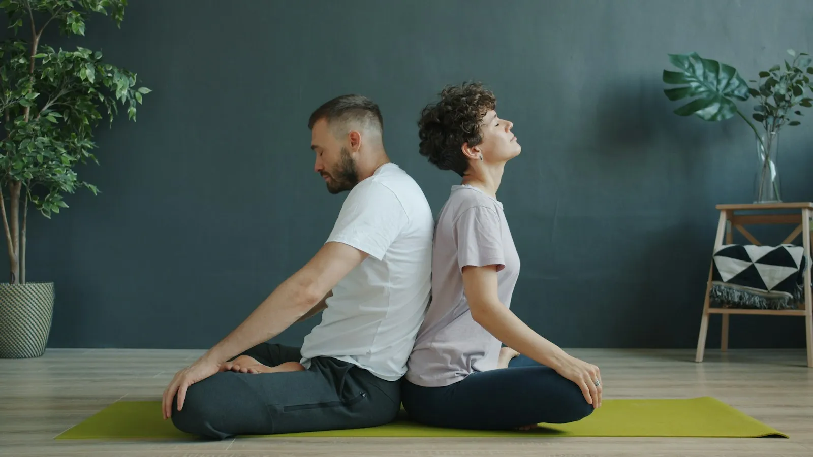 Ein Mann und eine Frau sitzen im Schneidersitz Rücken an Rücken auf einer gelbgrünen Yogamatte, die Hände auf den Knien, sie hebt den Kopf leicht, im Hintergrund eine dunkle Wand und Zimmerpflanzen.