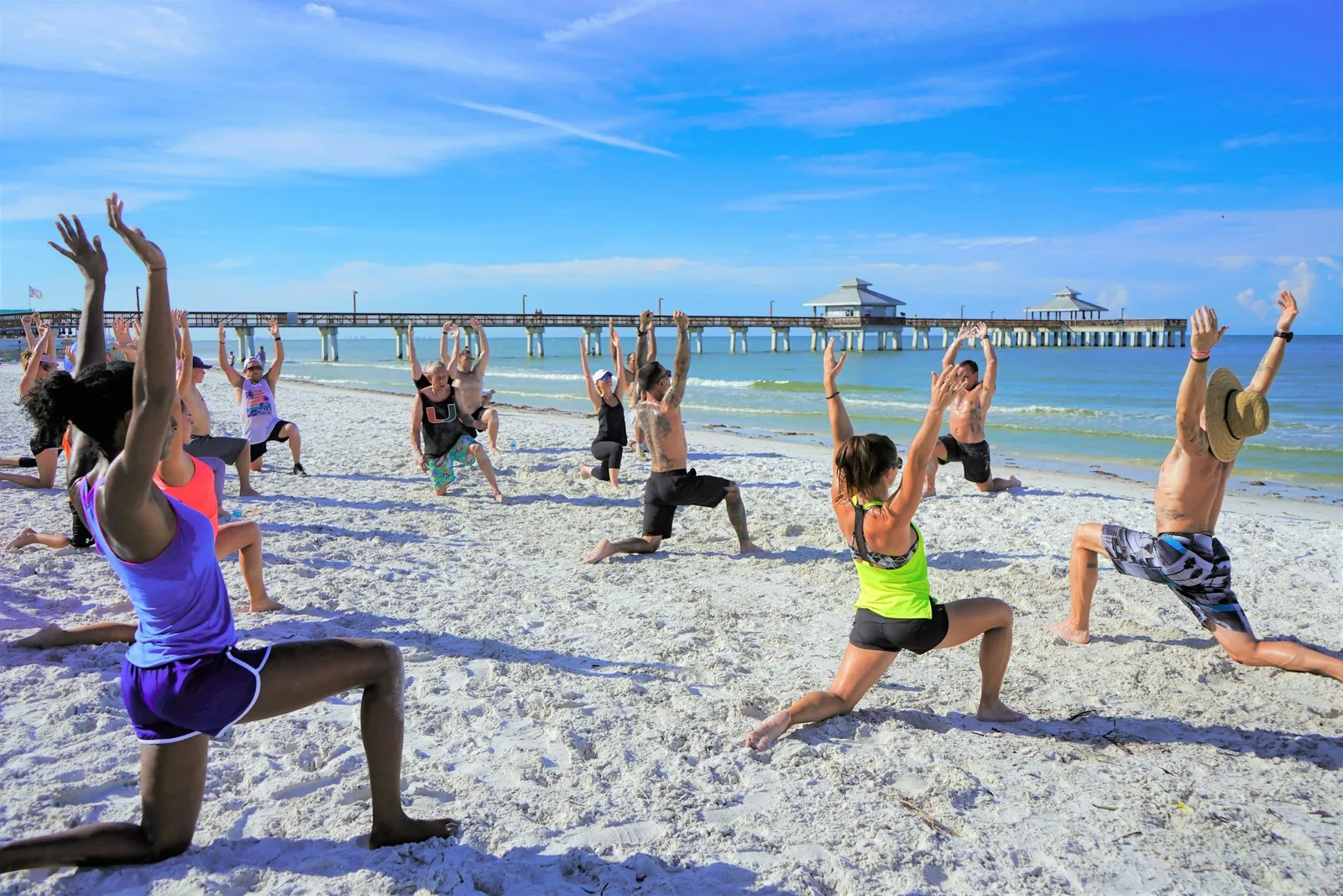 Große Gruppe von Menschen in der Krieger-Yoga-Pose an einem weißen Sandstrand mit Pier