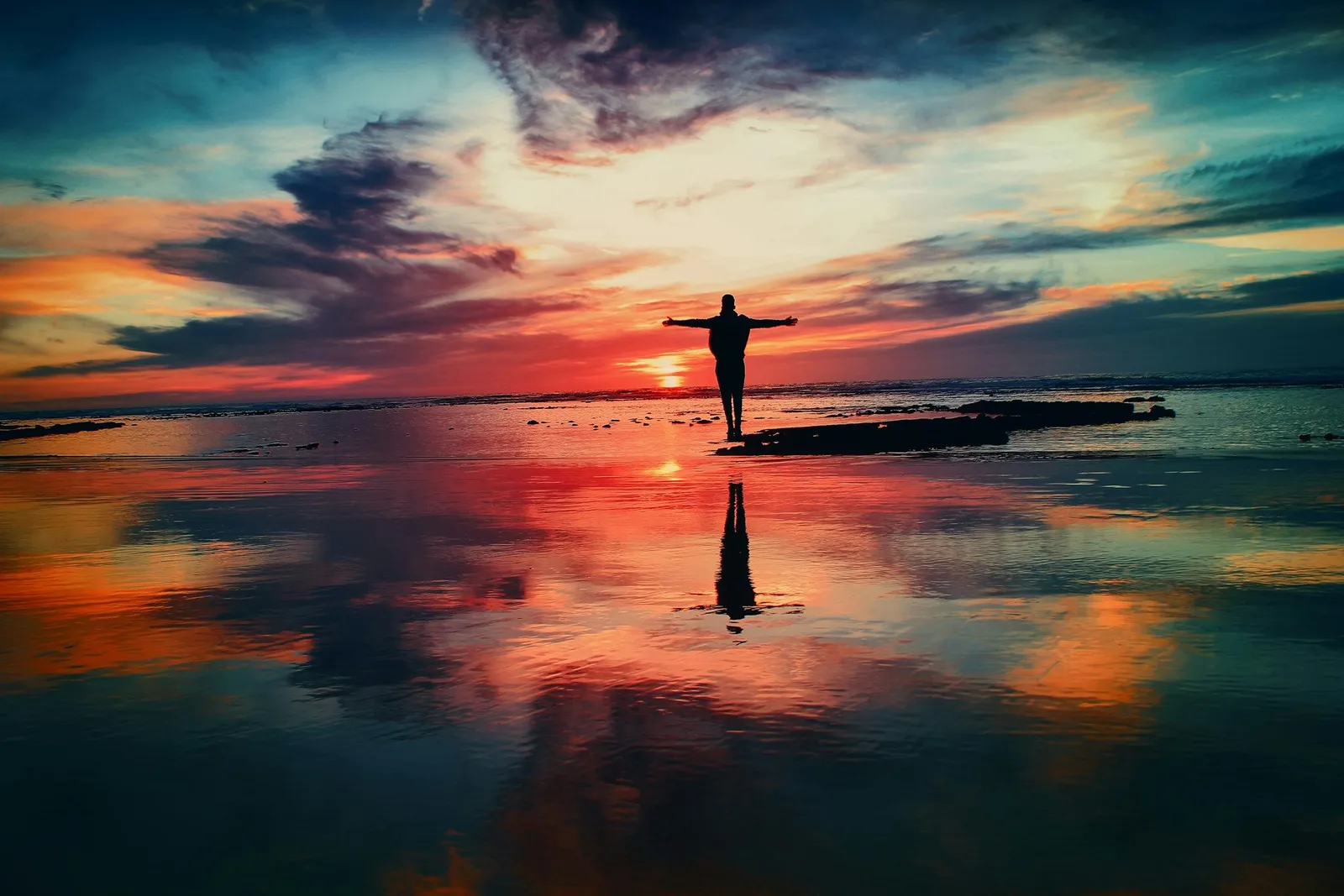 Silhouette einer Person auf nassem Sand bei Sonnenuntergang, Arme weit ausgestreckt, der leuchtende Abendhimmel spiegelt sich im flachen Wasser darunter.