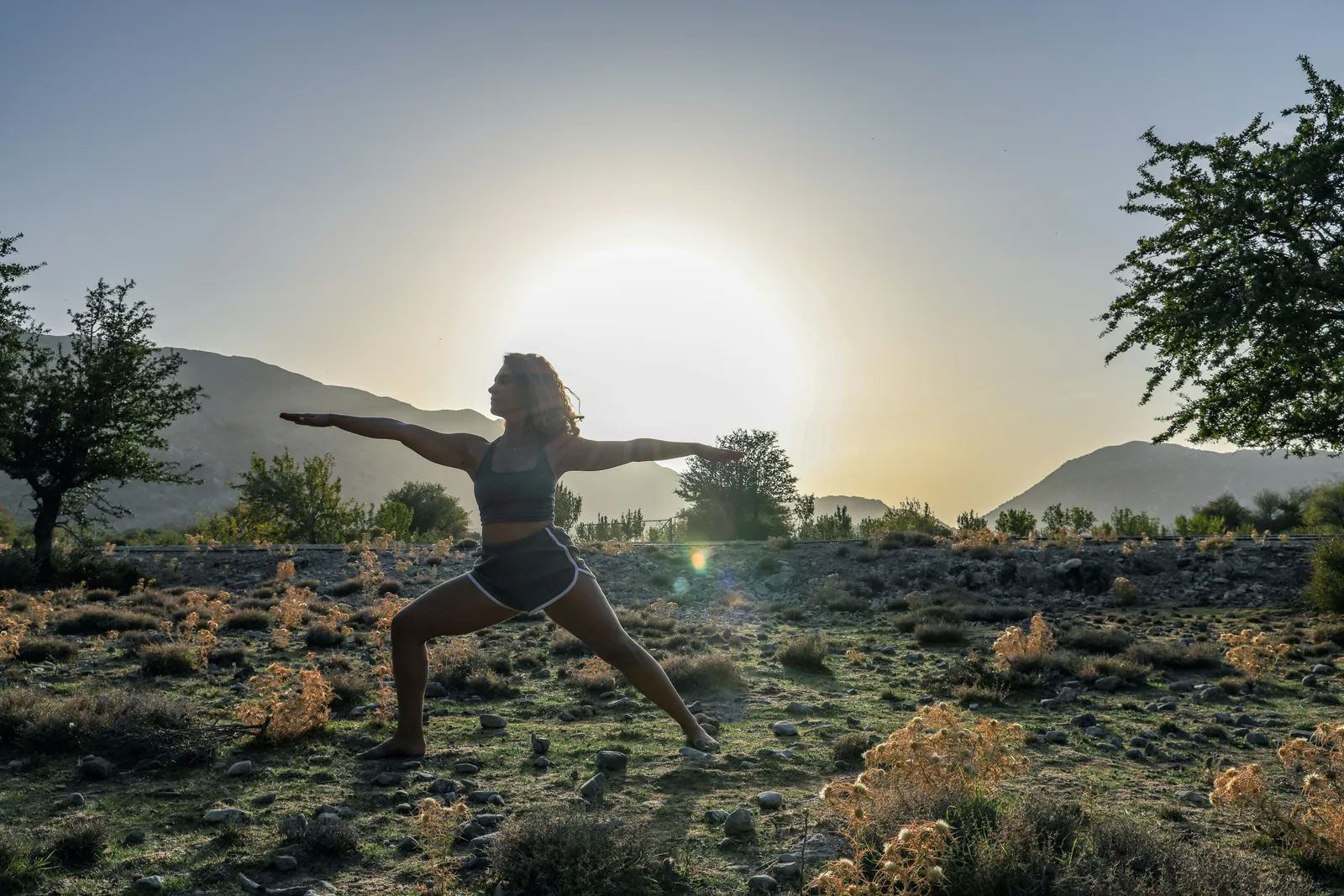 Frau in Krieger-II-Pose im Freien bei Sonnenuntergang, Berge im Hintergrund.