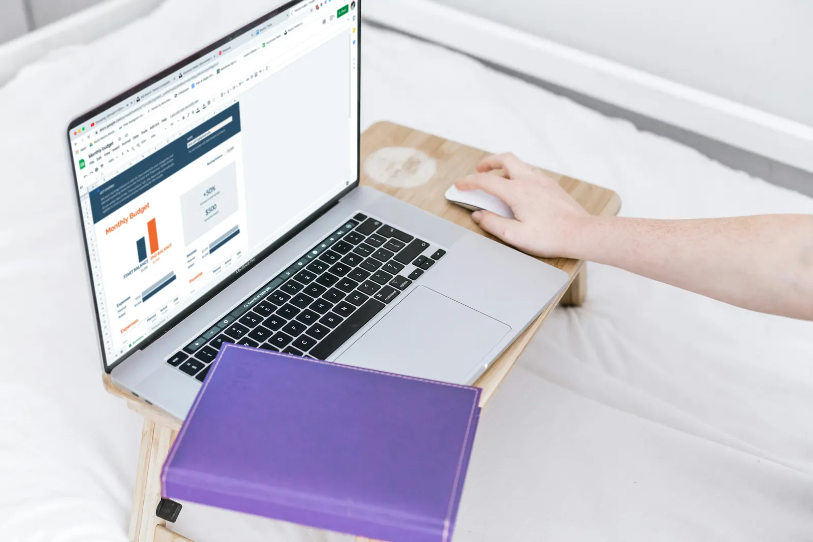 Person uses a laptop on a wooden bed-tray showing a Google Sheets monthly budget, with a purple notebook on top and white bedding around. Budget-planning scene.