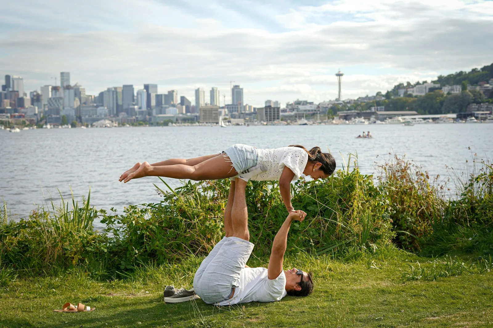 Paar macht Acroyoga auf Gras mit Stadtkulisse und See im Hintergrund, der Mann liegt auf dem Rücken und balanciert seine Partnerin in Plank-Position über sich.