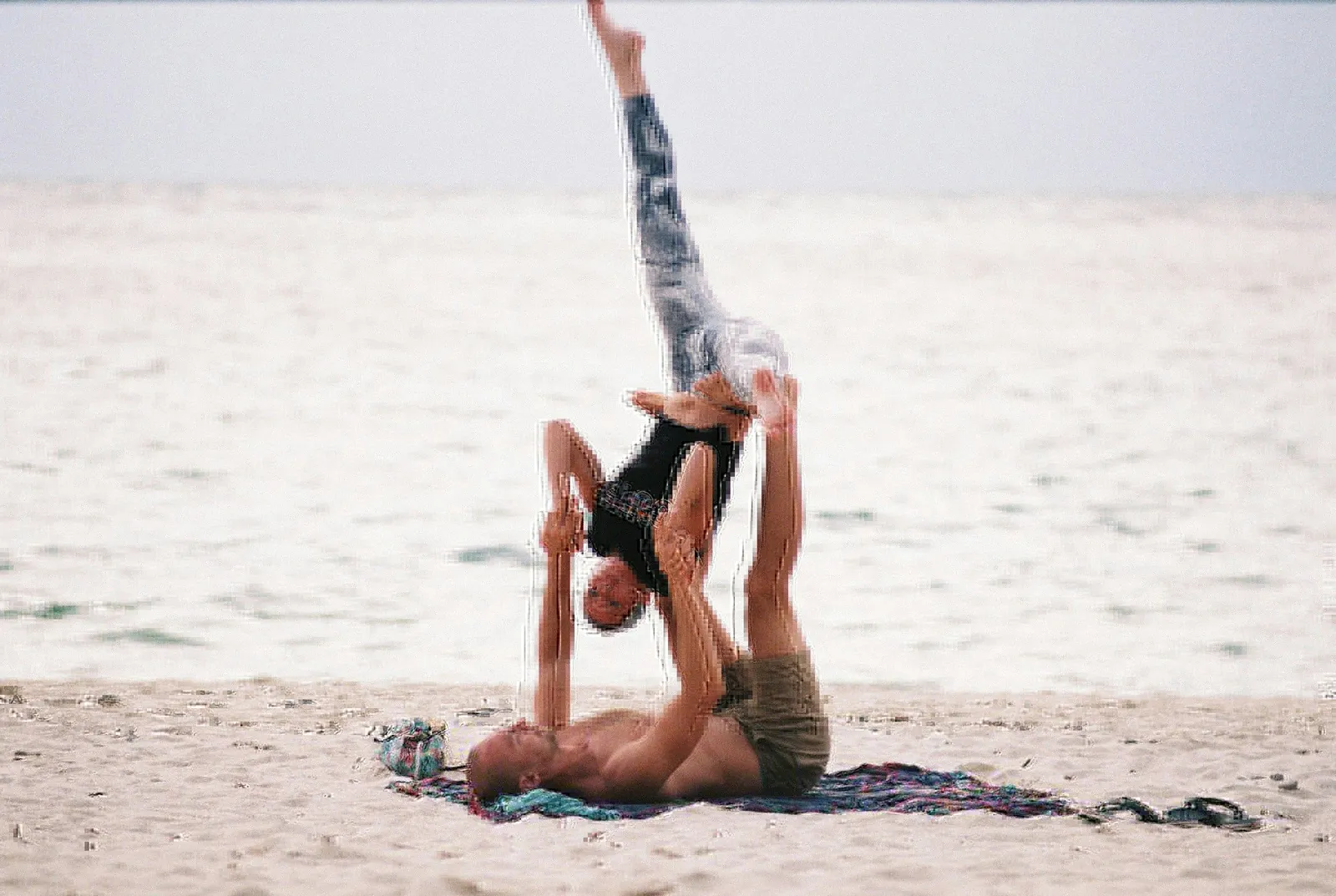 Paar beim Strand-Acroyoga, eine Person balanciert kopfüber auf den Füßen der anderen am Wasserrand.