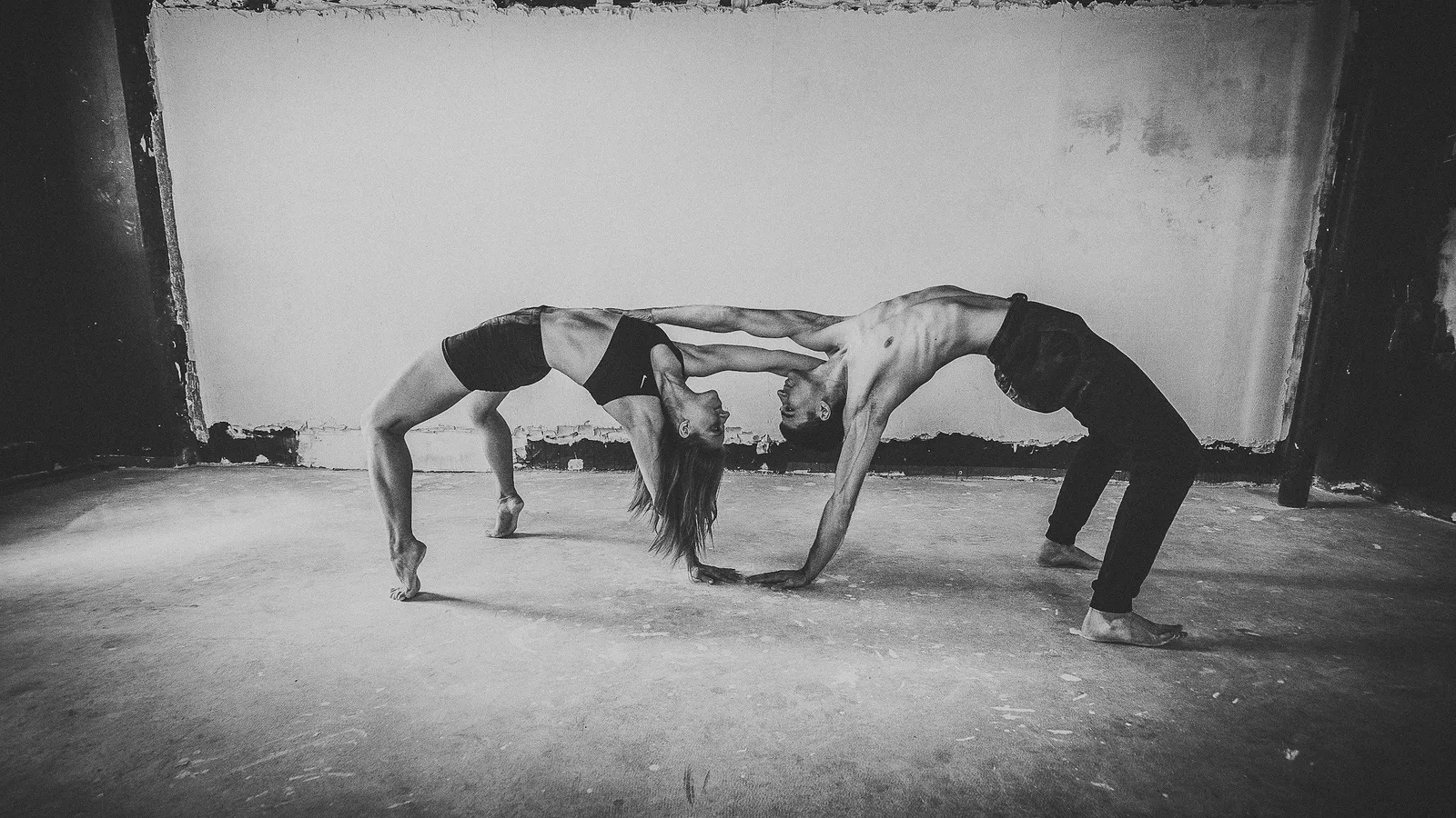 Schwarz-Weiß-Aufnahme eines Paares beim Partner-Acroyoga in ineinandergreifenden Rad-Posen in einem schmucklosen Betonraum.