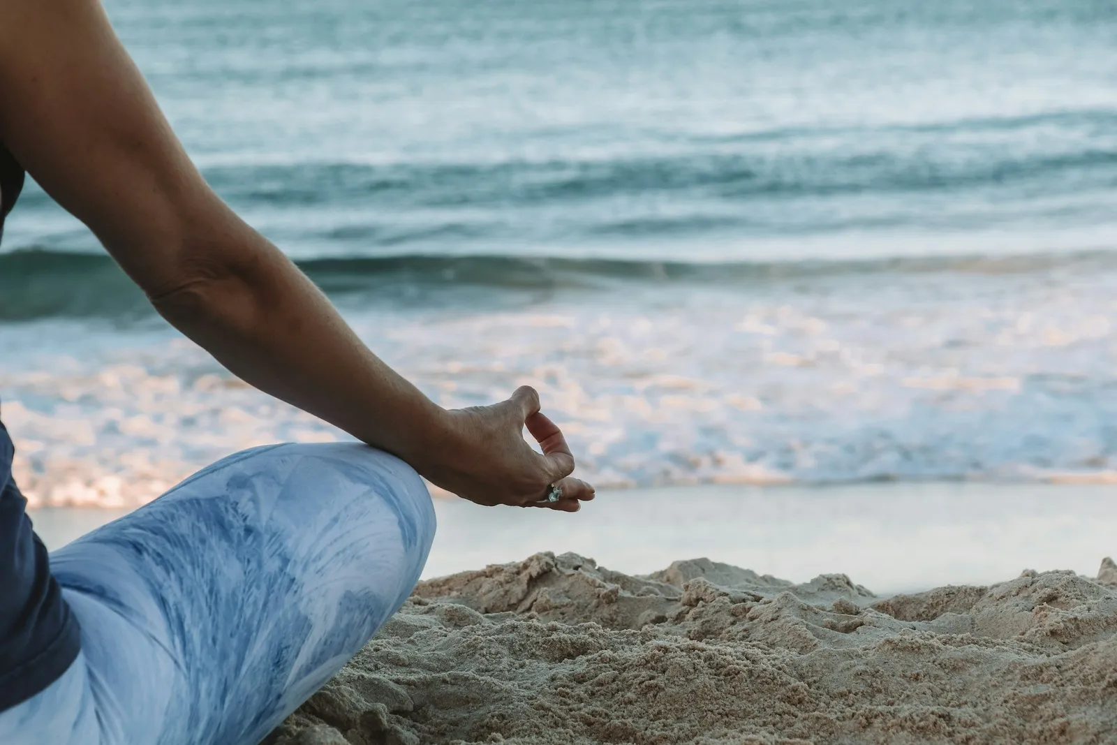Nahaufnahme einer Frau in blauen Leggings in Lotus-Pose auf Sand mit einer Hand in Chin-Mudra, Meereswellen dahinter.
