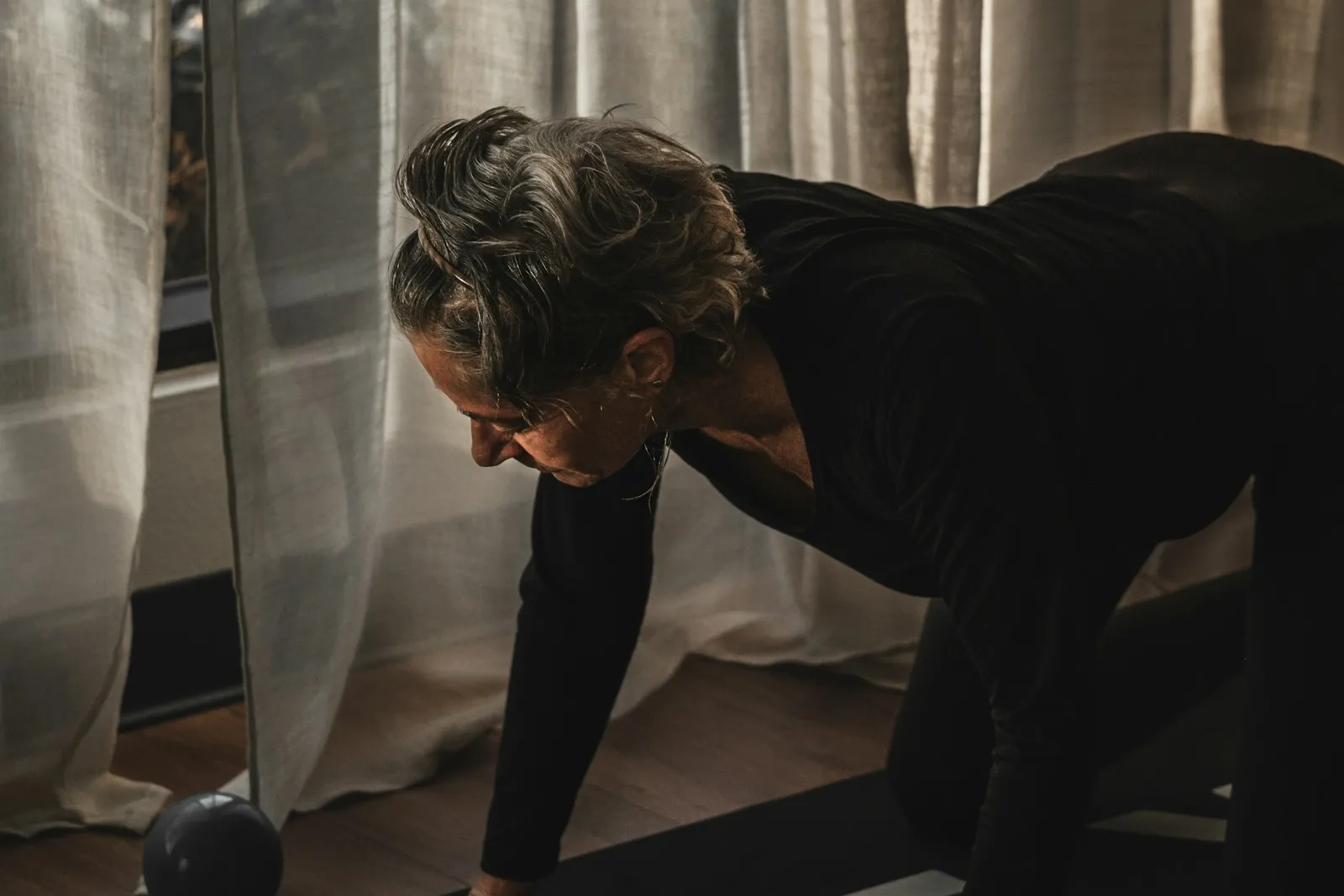 Eine Frau mit grau meliertem, hochgestecktem Haar geht im Halbdunkel auf der Yogamatte in den Vierfüßlerstand, den Blick nach unten. Hinter ihr fällt gedämpftes Licht durch helle Leinenvorhänge, neben ihr liegt ein kleiner Gymnastikball.
