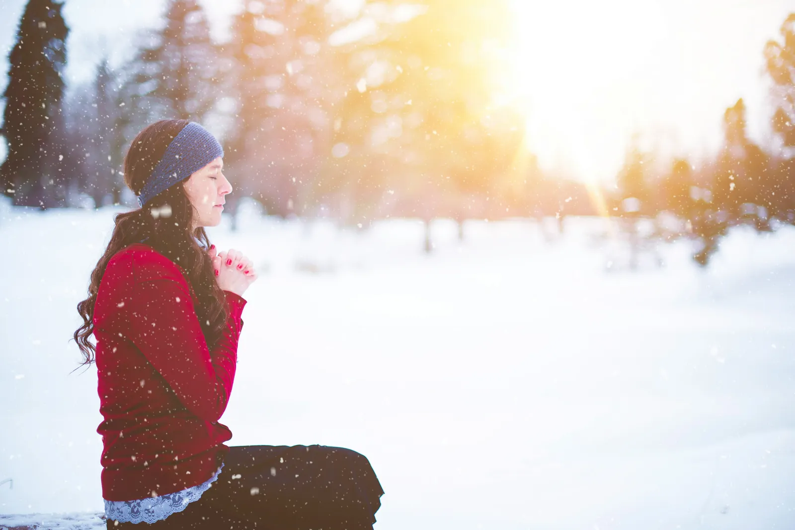 Frau in rotem Pullover in Gebetshaltung in einer verschneiten Lichtung, warmes Sonnenlicht bricht durch die Bäume.